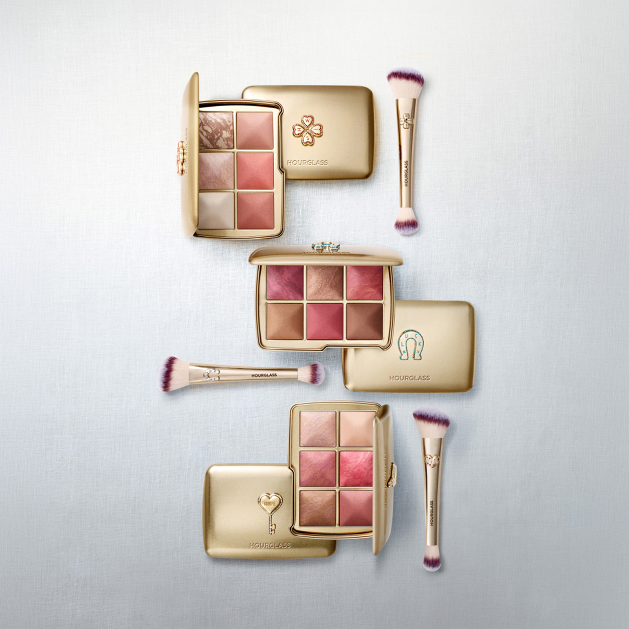 Hourglass makeup collection featuring eyeshadow palettes in warm pink and brown tones, powder compacts with gold detailing, and dual-ended makeup brushes with purple bristles