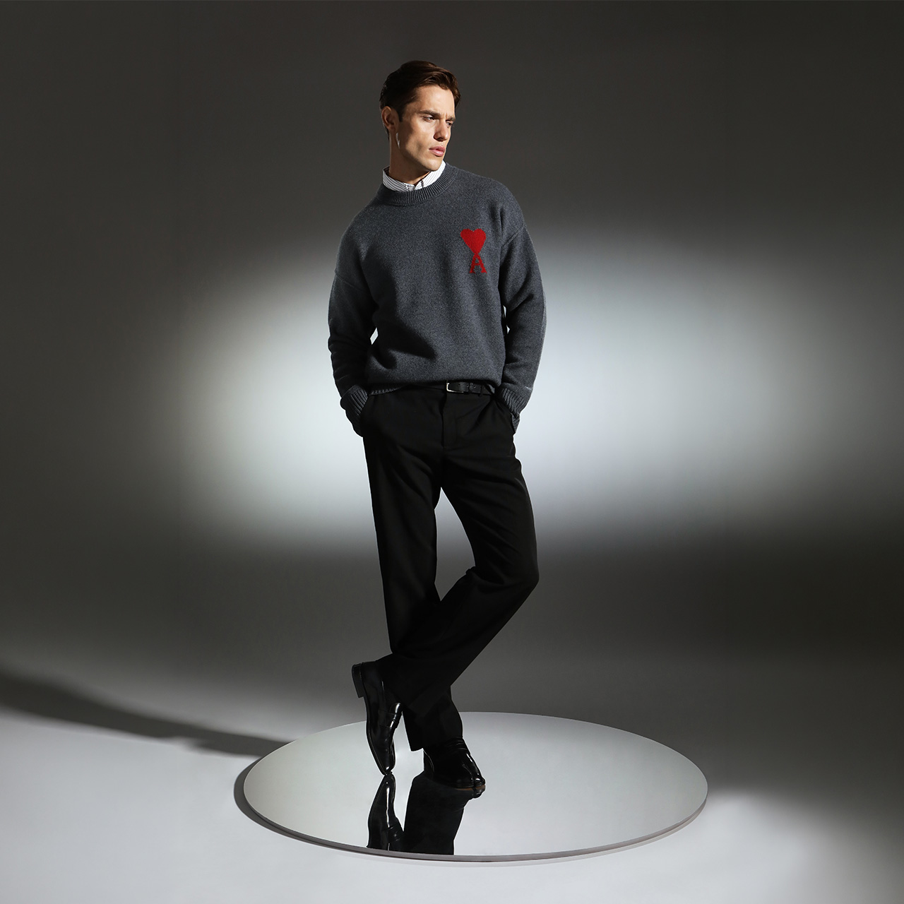 Man wearing charcoal sweatshirt with red emblem and black trousers standing on circular platform against grey background