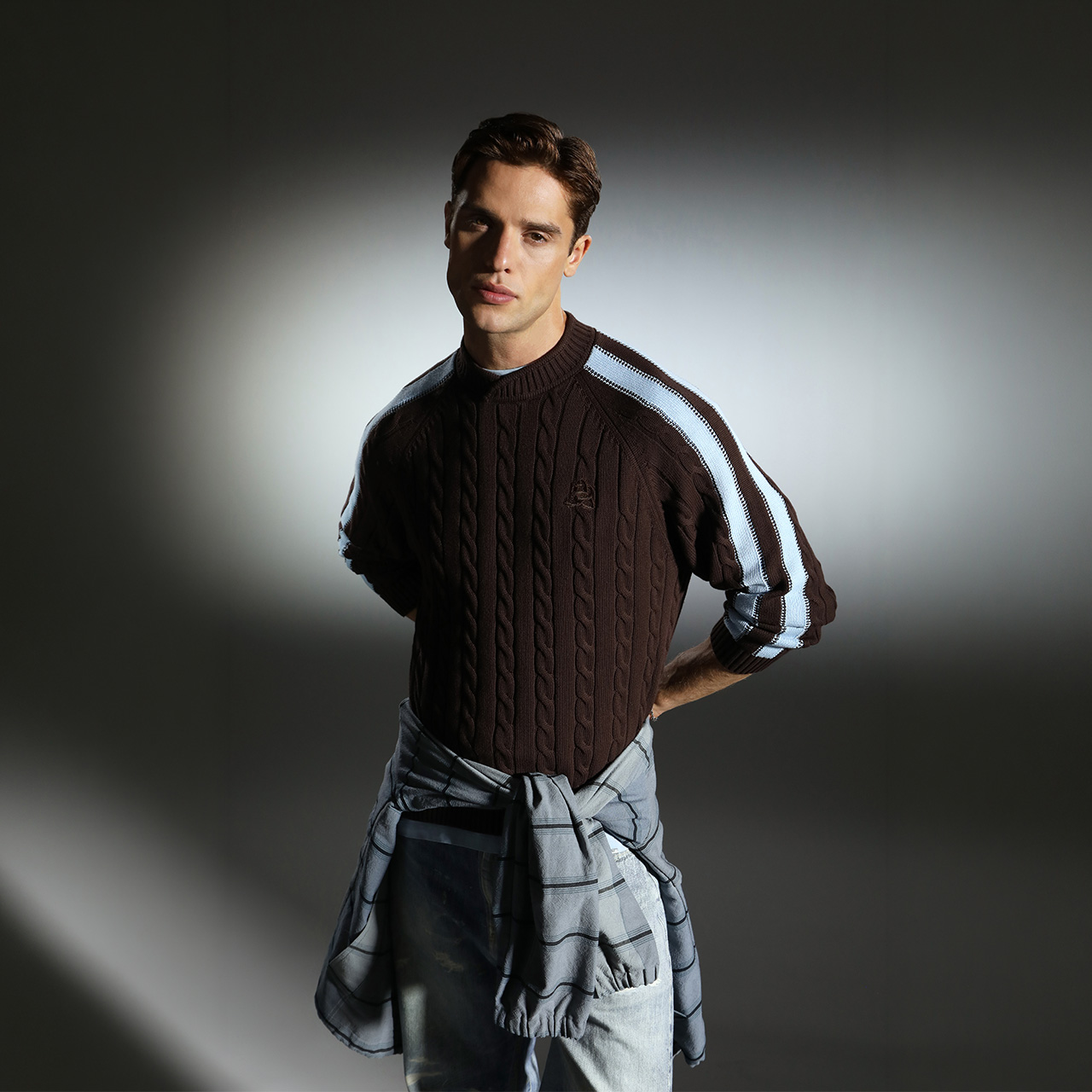 Man wearing black cable-knit jumper with white stripe detail and light blue trousers against neutral background