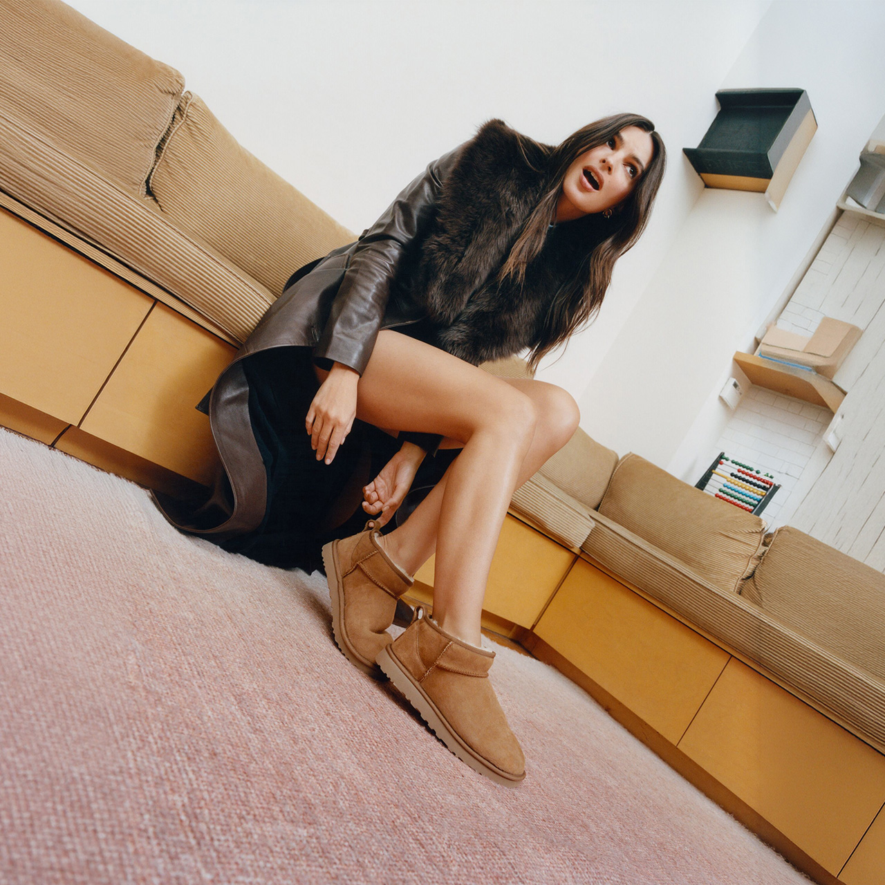Woman wearing camel suede ankle boots and brown leather jacket sitting on yellow bench in modern interior