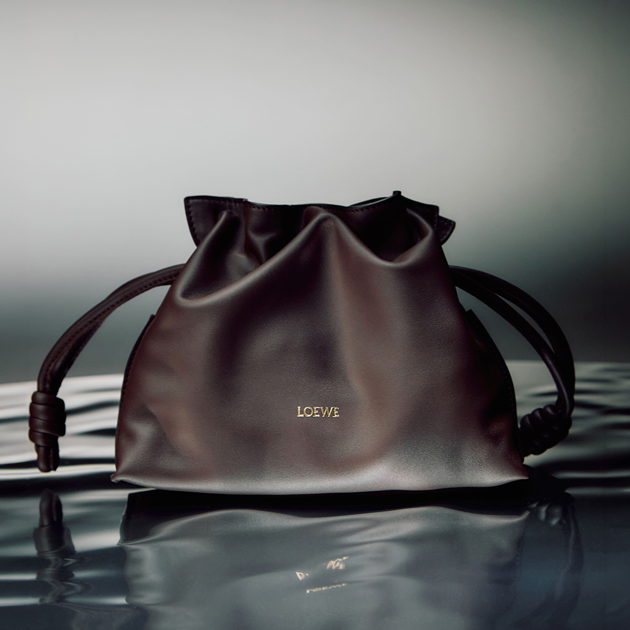 Burgundy leather drawstring bucket bag with gold branding displayed on reflective water surface