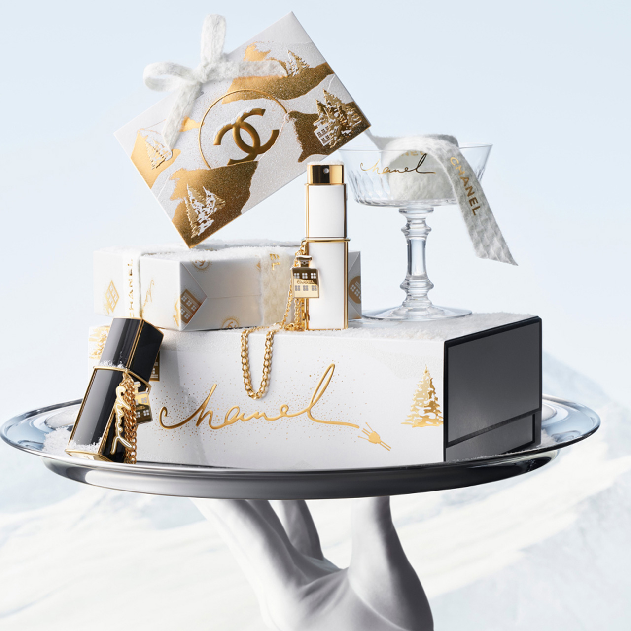 Stylish holiday-themed Chanel products on a silver tray, including wrapped gifts, jewelry, and perfume bottles, with a snowy backdrop.