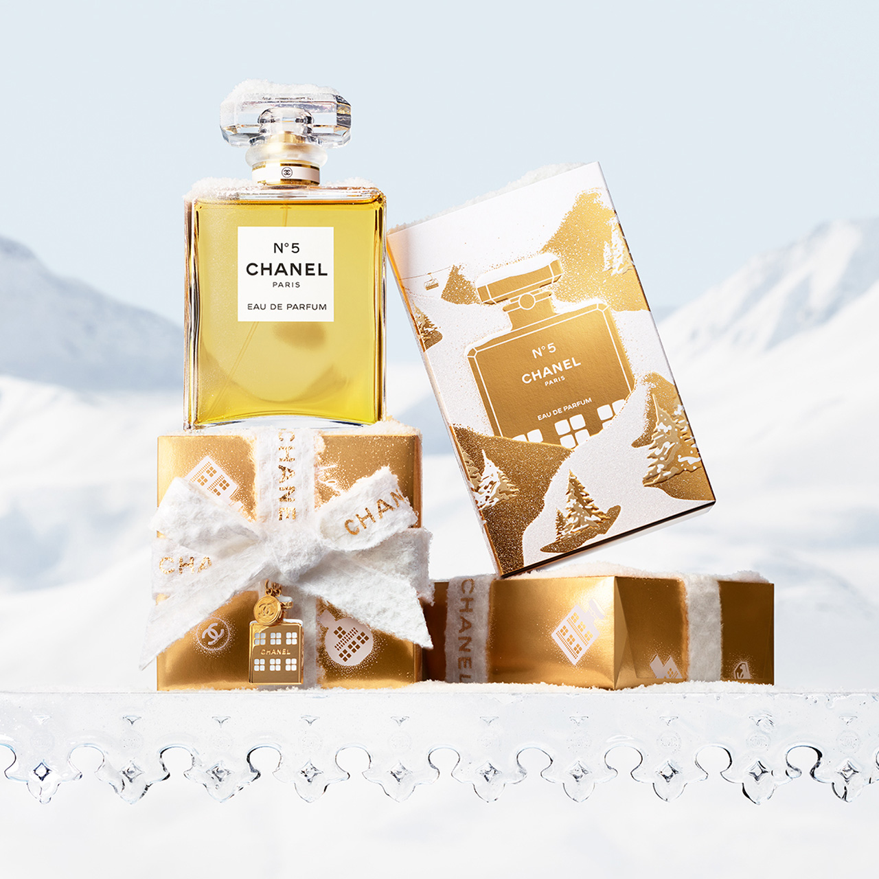 Chanel No. 5 eau de parfum bottle in yellow glass with crystal stopper, displayed with festive white and gold holiday gift boxes and greeting card
