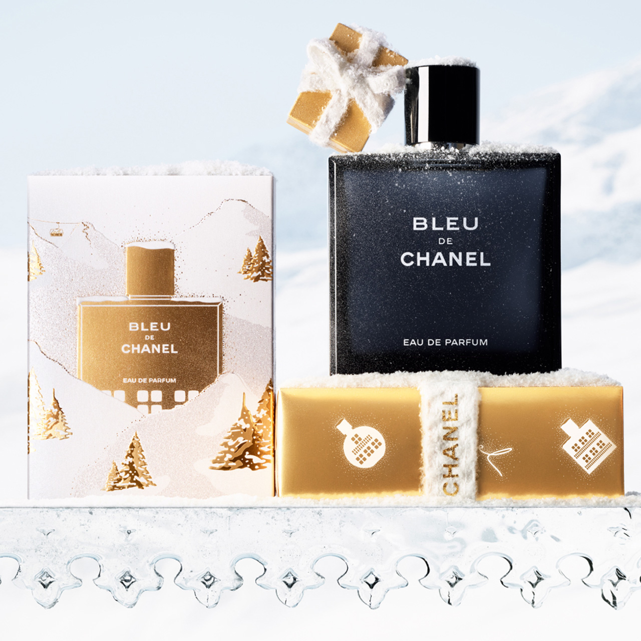 Bleu de Chanel eau de parfum in black bottle with white text, surrounded by festive gift boxes and white holiday decorations on snowy surface