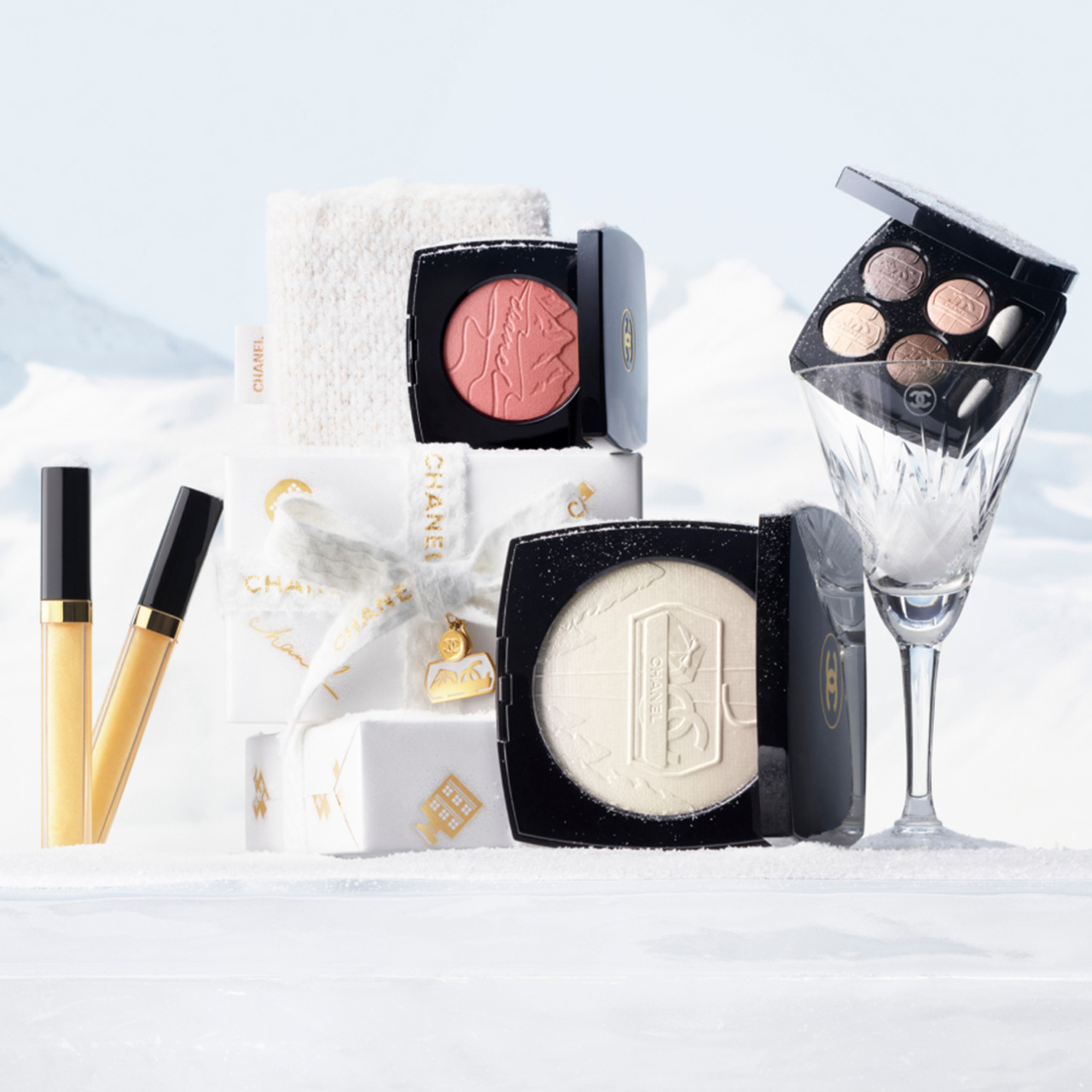 Collection of Chanel makeup products including lipgloss, eyeshadow palette, powder and rouge, arranged with holiday gift boxes and crystal glass