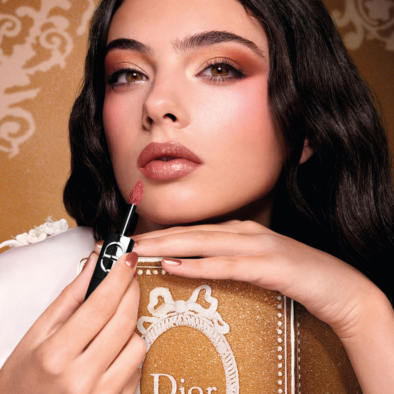 A woman with makeup holds a lipstick applicator near her lips, resting her hand on a Dior-branded book against a patterned background.