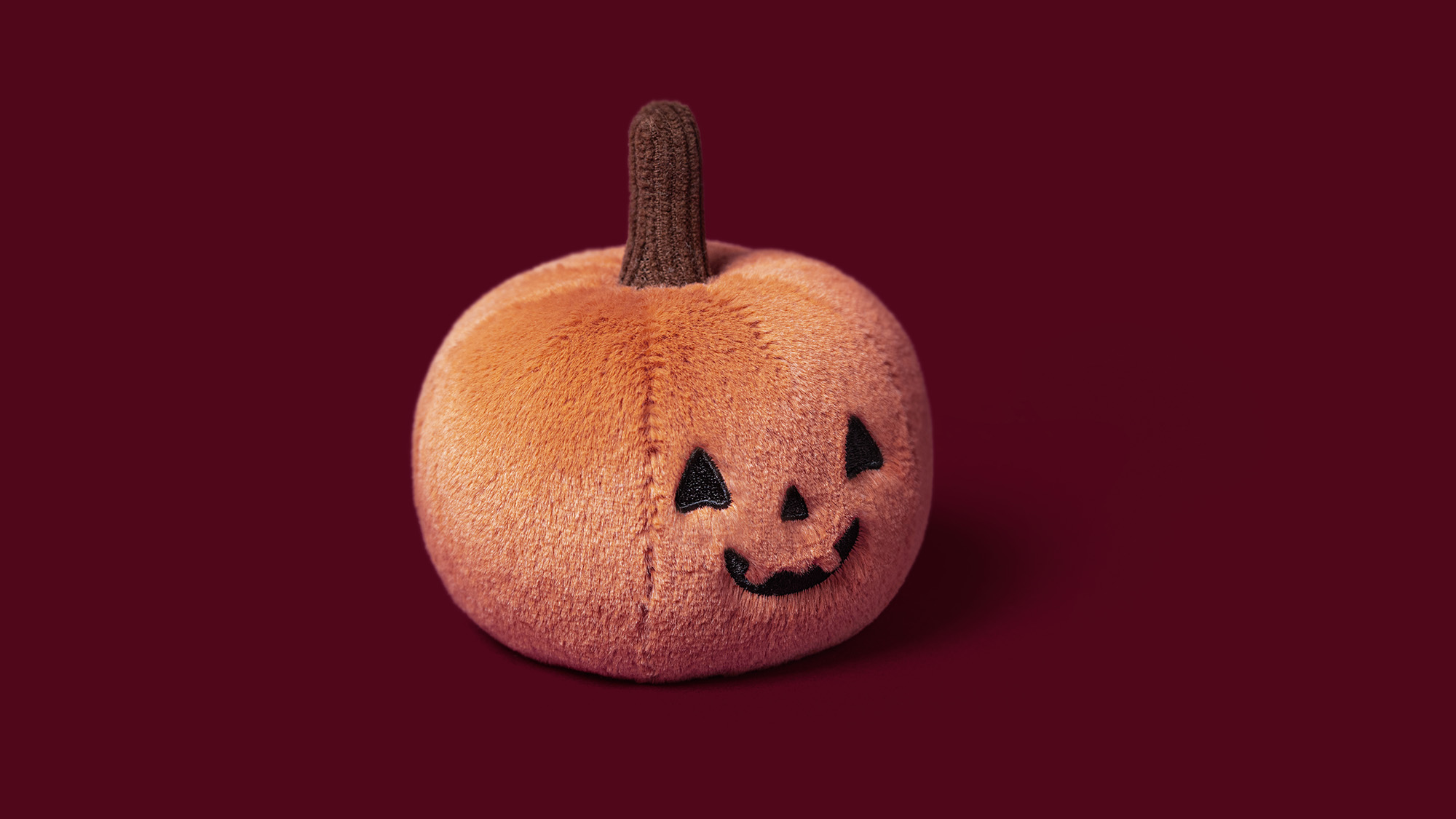 Soft plush pumpkin toy with jack-o'-lantern face and brown stem against deep burgundy background