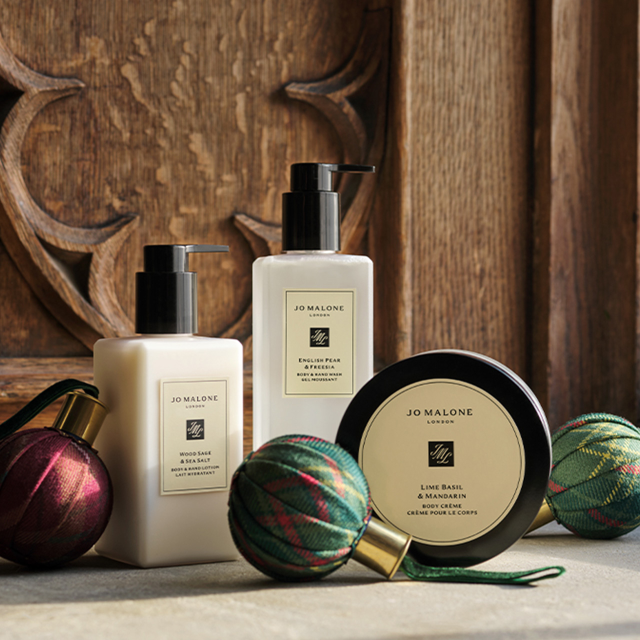 Jo Malone London bath and body products including body cr&egrave;me, bath oil, and body wash displayed with festive ornaments against wooden backdrop