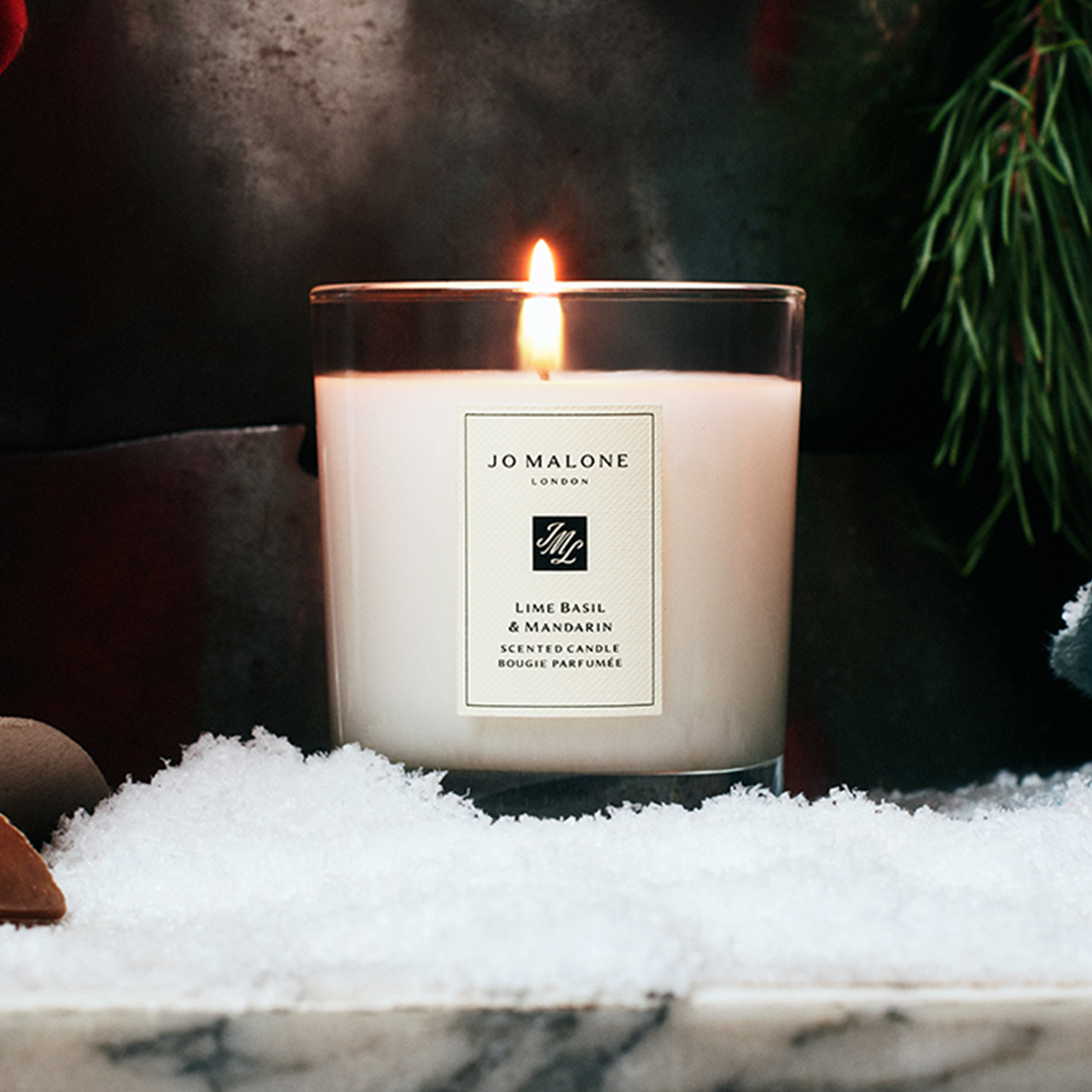 Lit scented candle with Lime Basil & Mandarin label surrounded by white snow, evergreen sprigs, and festive decorations.