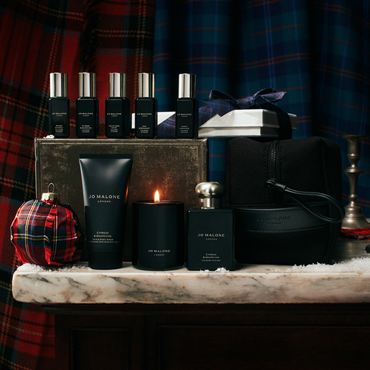 Collection of luxury fragrance bottles, candles, and black leather accessories displayed on marble shelf with tartan backdrop.