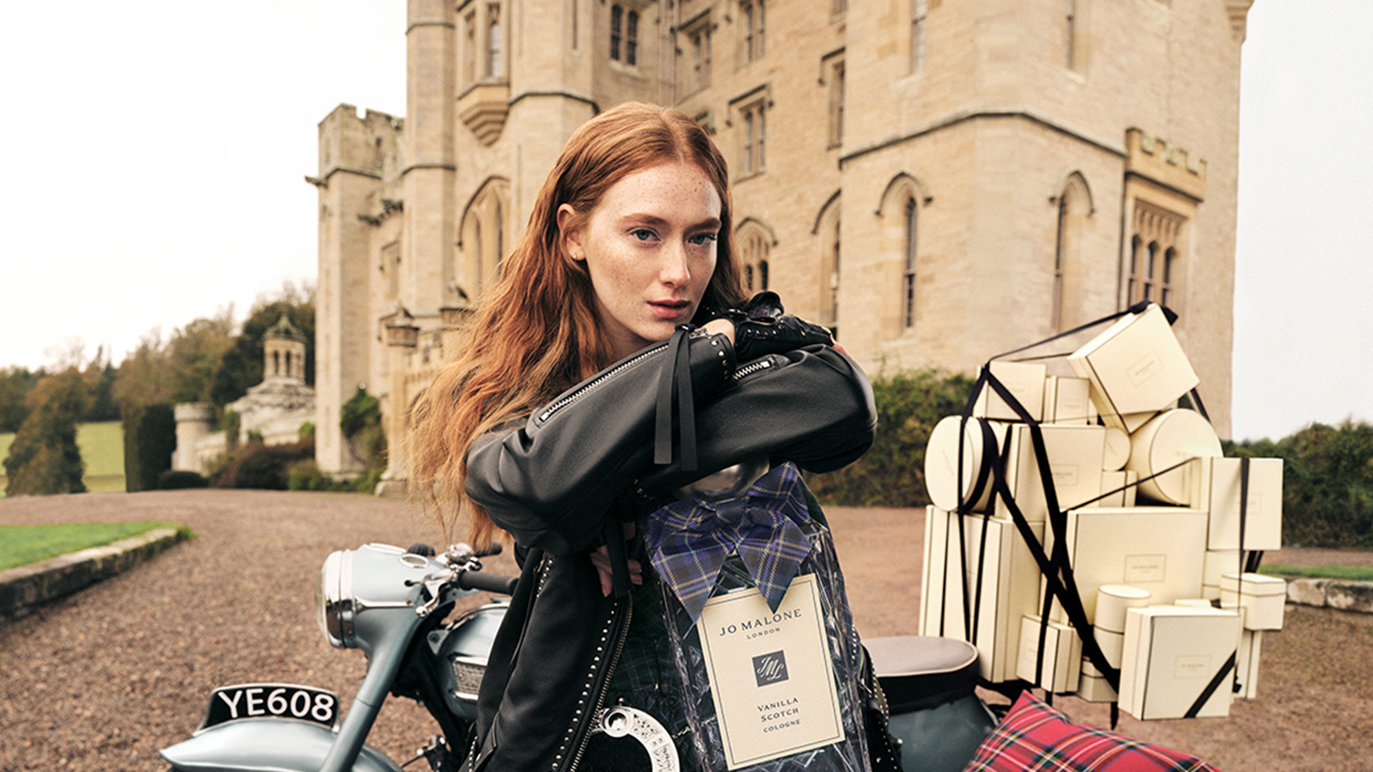 Woman on motorcycle wearing black leather jacket with Jo Malone London fragrance bottle and gift boxes stacked beside her at castle estate