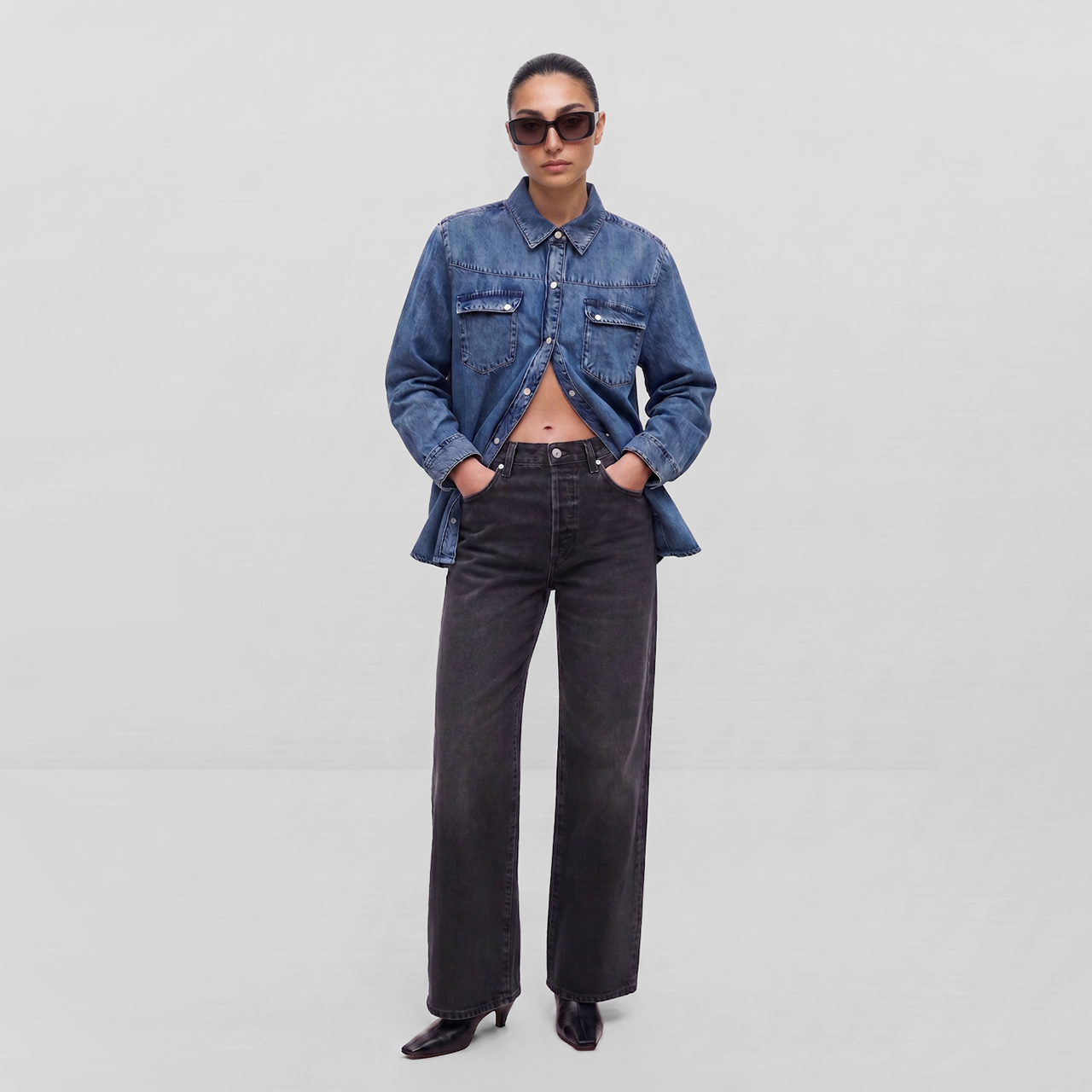 Woman wearing blue denim jacket, black jeans, and sunglasses standing against white background