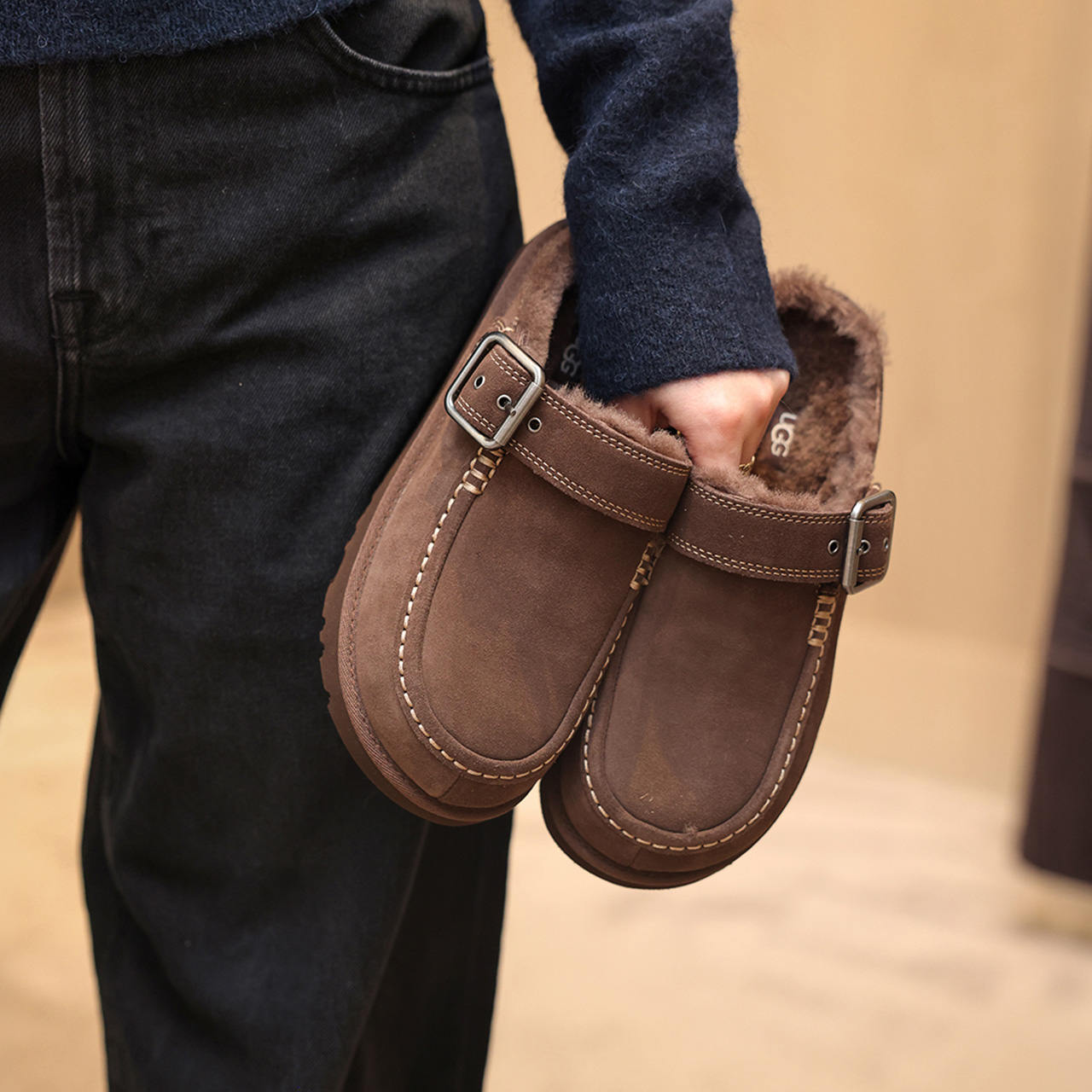 Woman wearing navy jeans and brown suede sheepskin-lined Ugg Evelina slippers with buckle detail