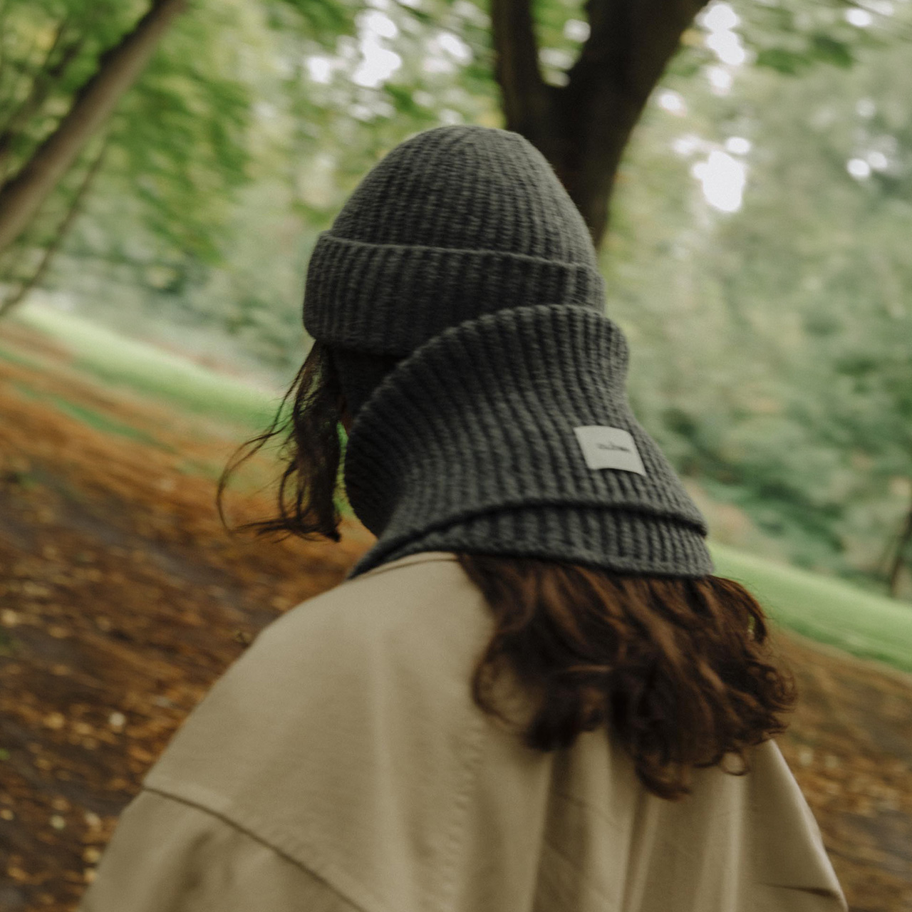 Woman from behind wearing black ribbed beanie and cream jacket on forest path with green foliage