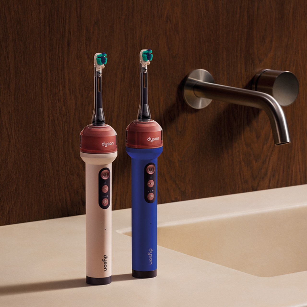 Two electric toothbrushes in cream and blue with coloured bands beside stainless steel bathroom tap over sink