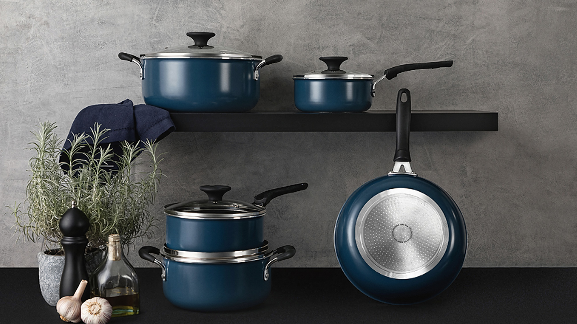 Navy blue cookware set displayed on black shelf with fresh herbs, garlic, and olive oil on grey background