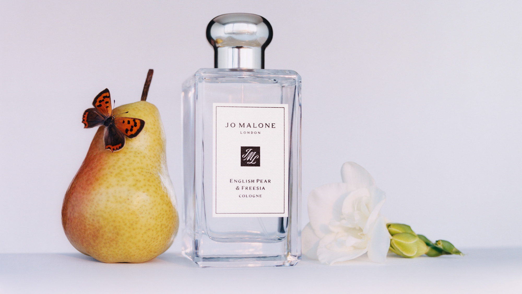 Perfume bottle beside a pear with a butterfly, and a white flower.