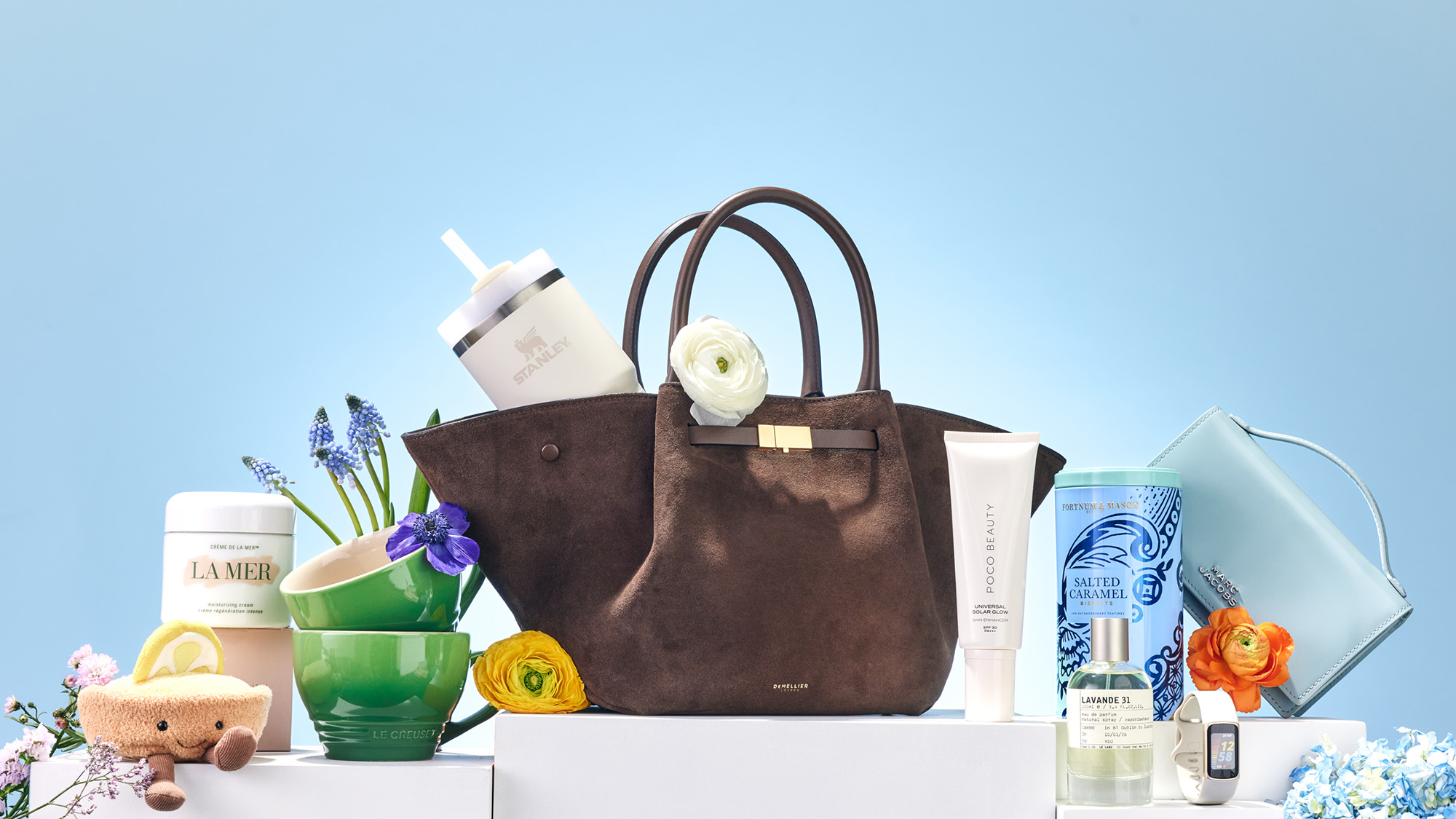 A brown handbag is surrounded by various beauty products, flowers, and a drink cup in a bright setting.