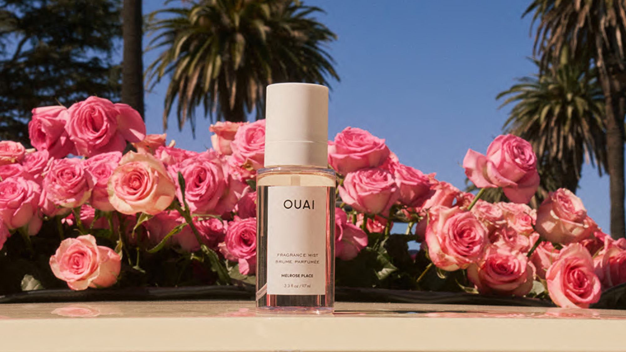 A perfume bottle on a surface surrounded by pink roses, with palm trees in the background.