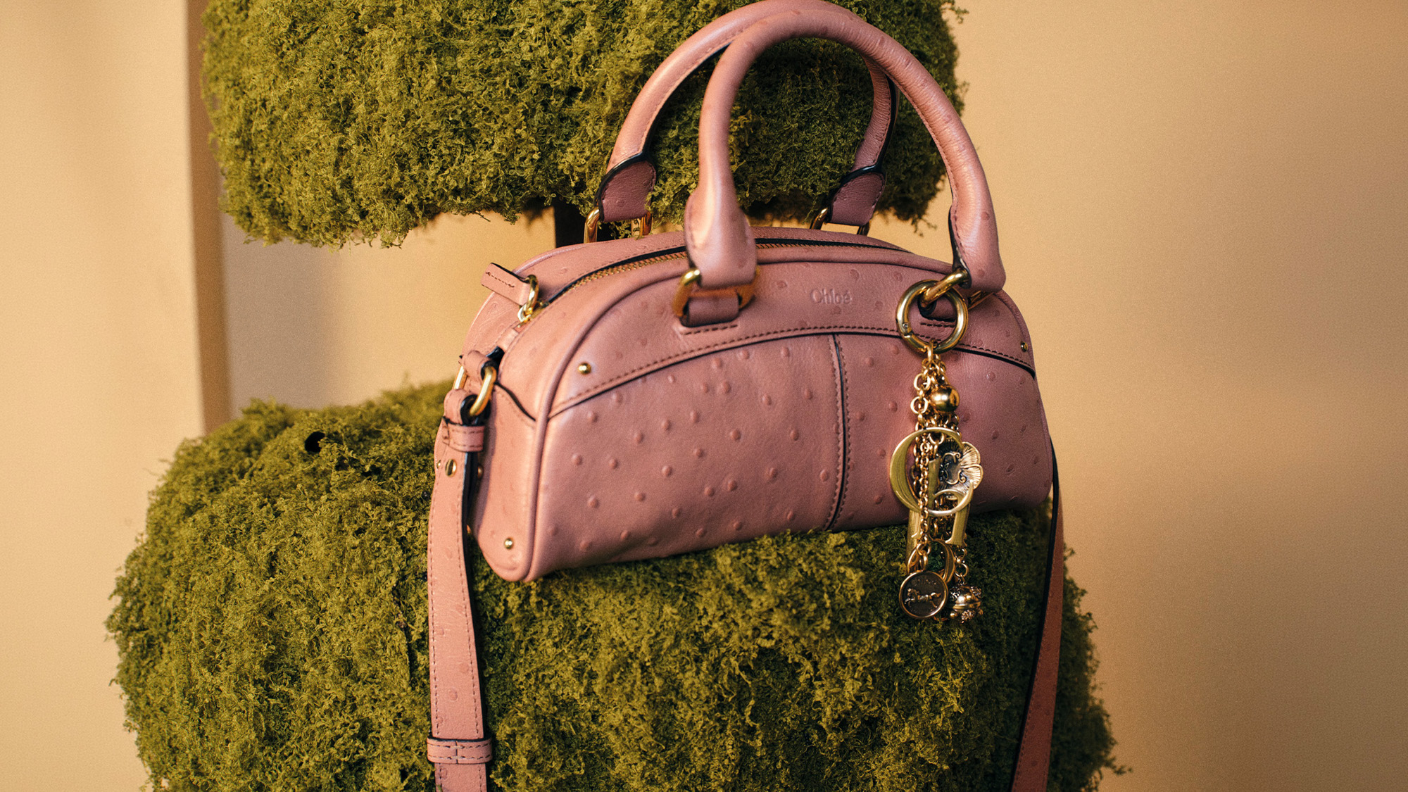 A pink handbag with a charm hangs on a moss-covered mannequin.