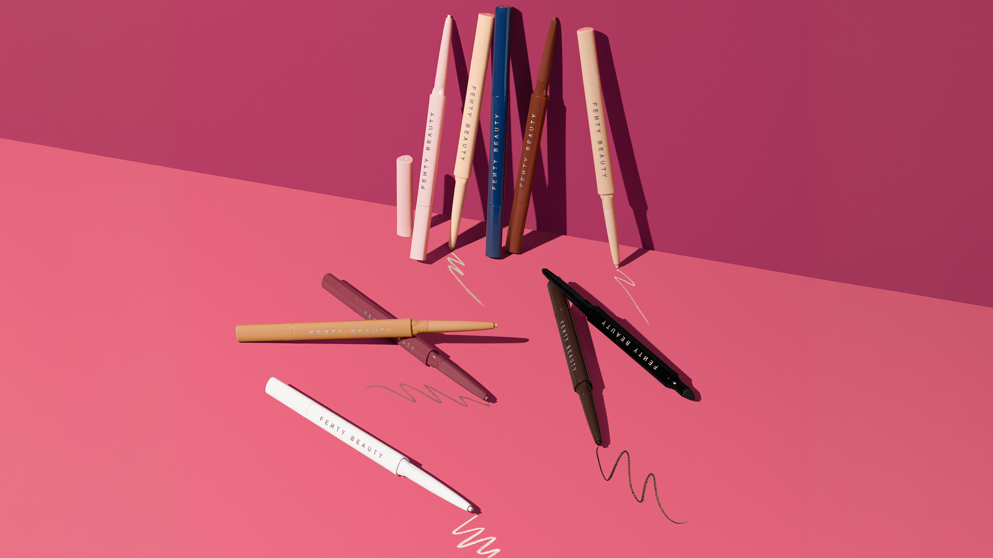 Assorted colored eyeliner pencils on a pink surface with scribbles beside them.