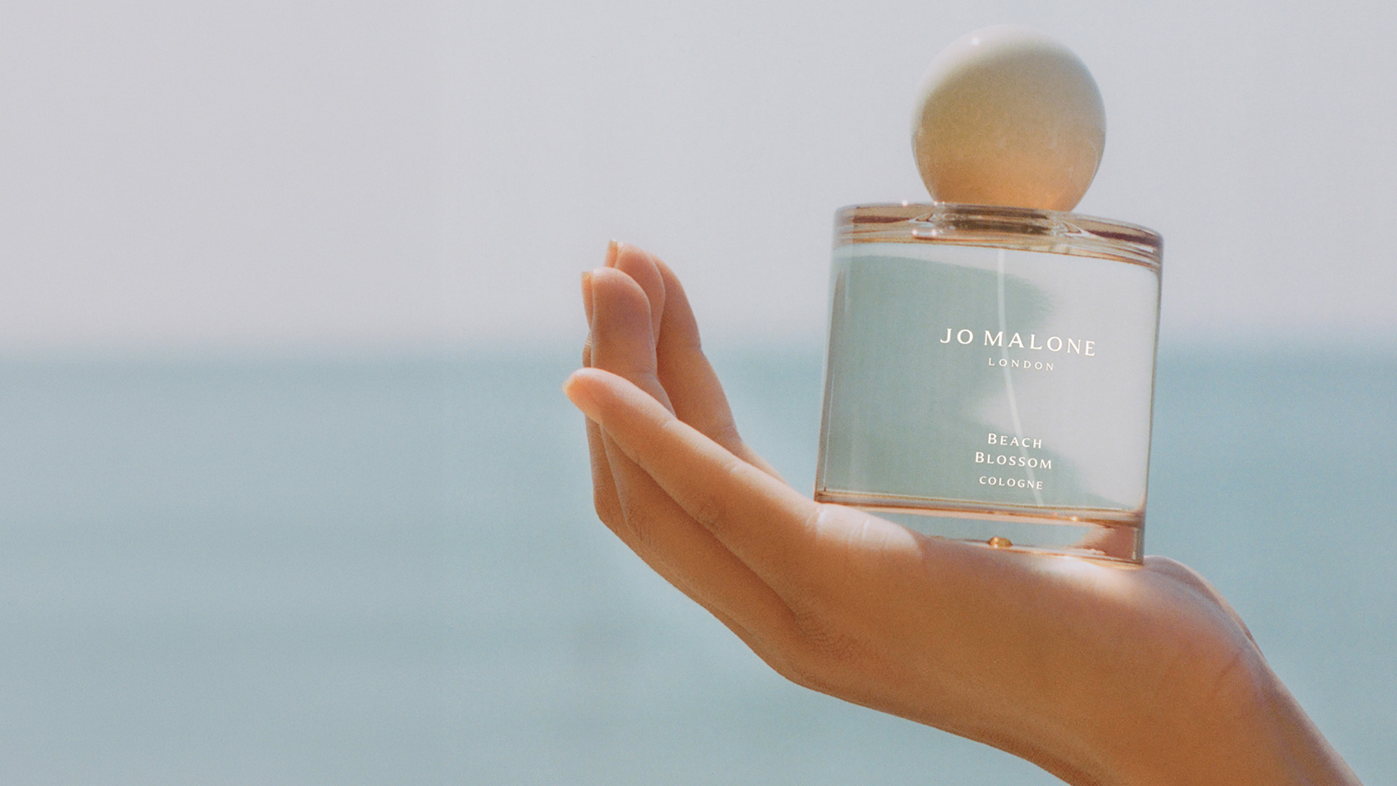 A hand holds a Jo Malone London perfume bottle against a blurred seascape background.