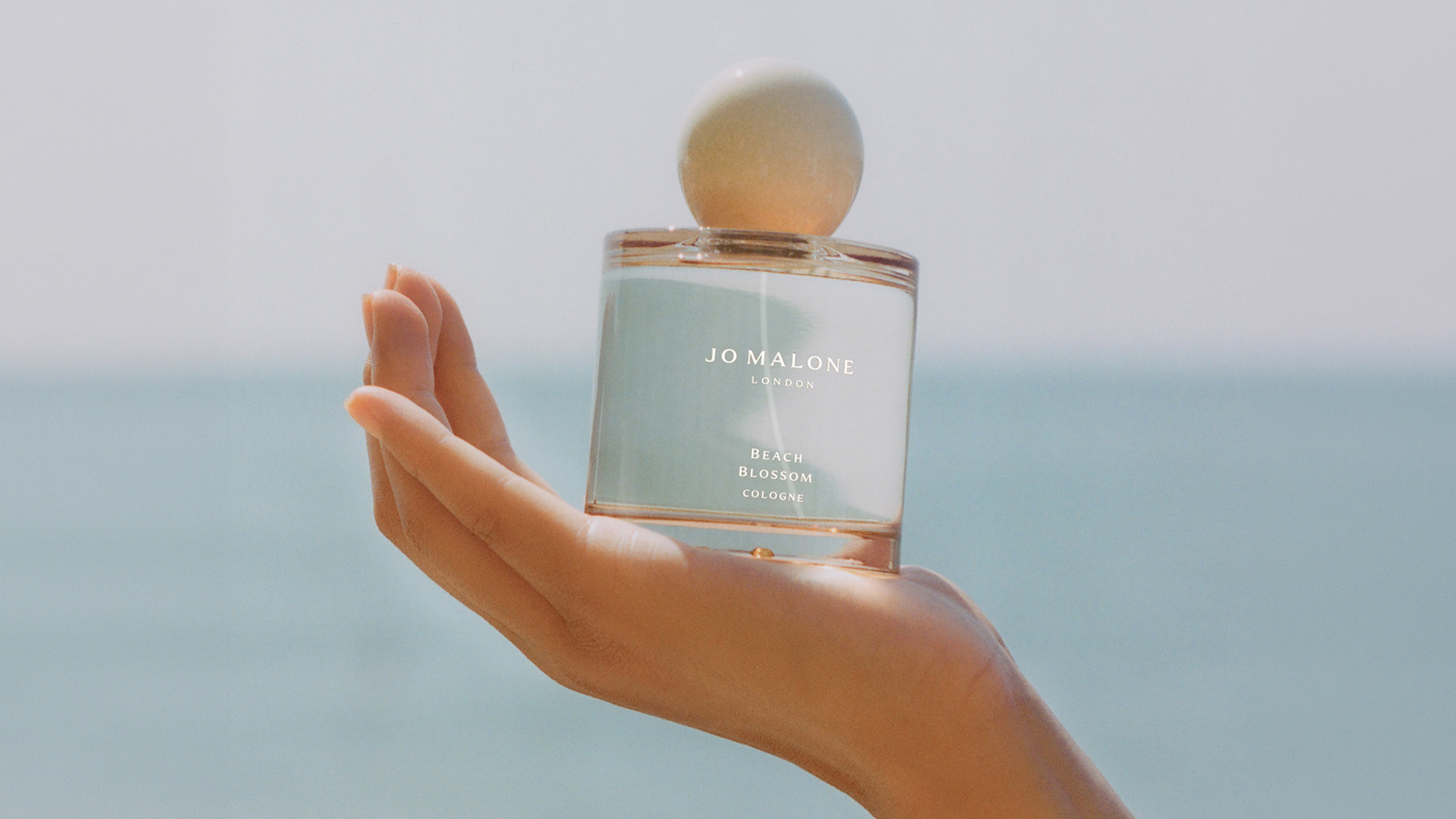 A hand holds a Jo Malone London perfume bottle against a blurred seascape background.