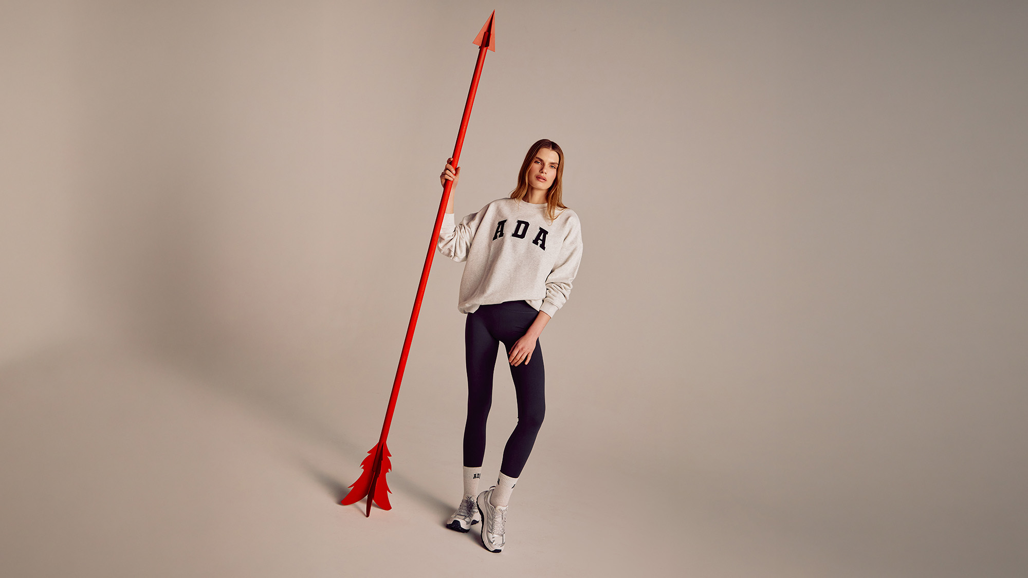 A woman stands holding a tall red arrow, wearing a white sweater and dark leggings with white boots.