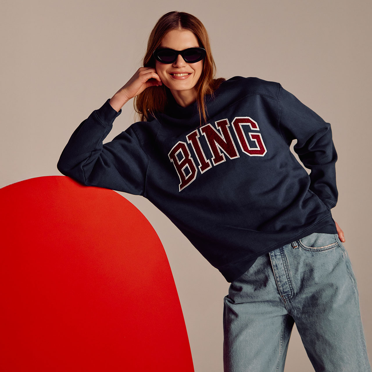 A person wearing sunglasses and a navy 'BING' sweatshirt leans against a red wall, smiling, with hands on hip and head.