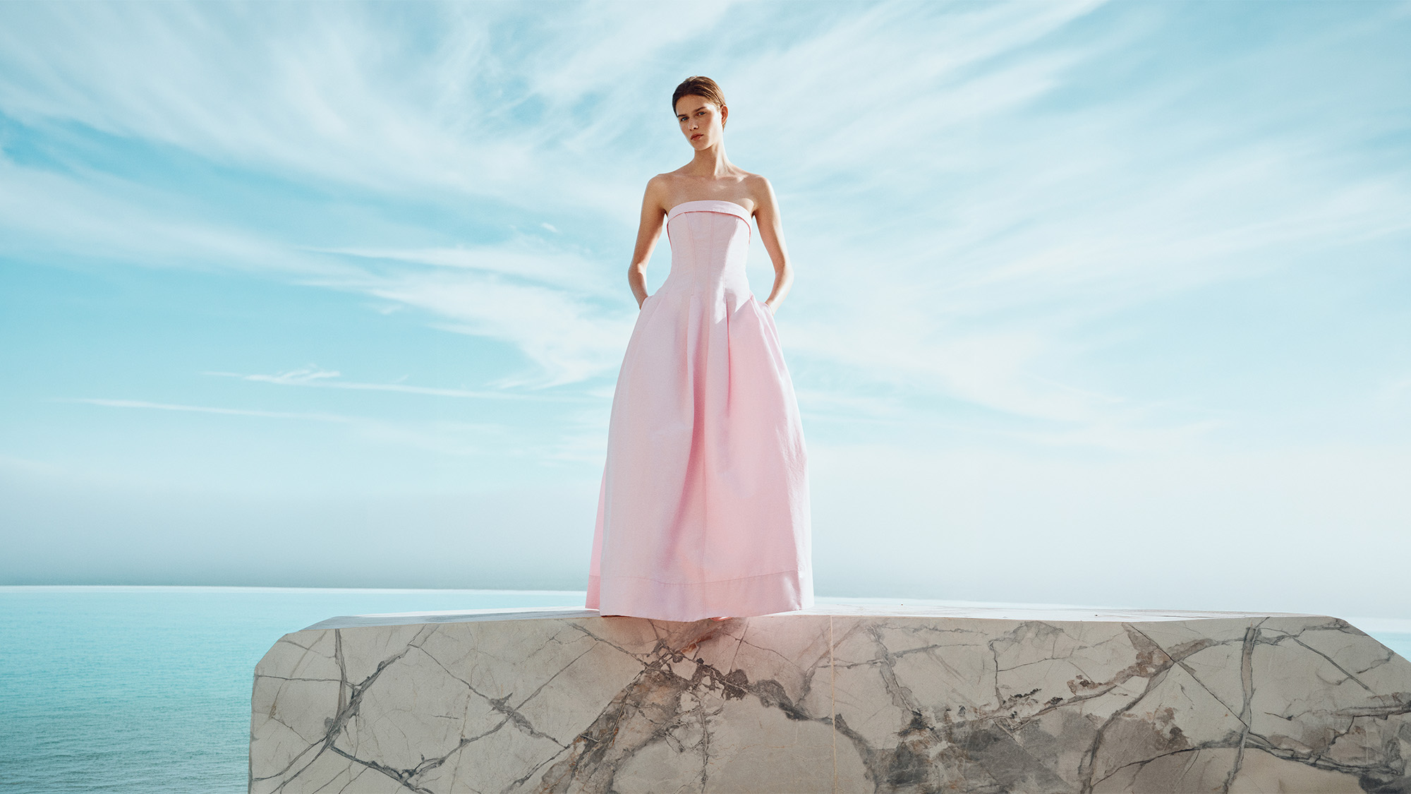 Woman in pale pink strapless ball gown standing on marble platform overlooking calm turquoise sea