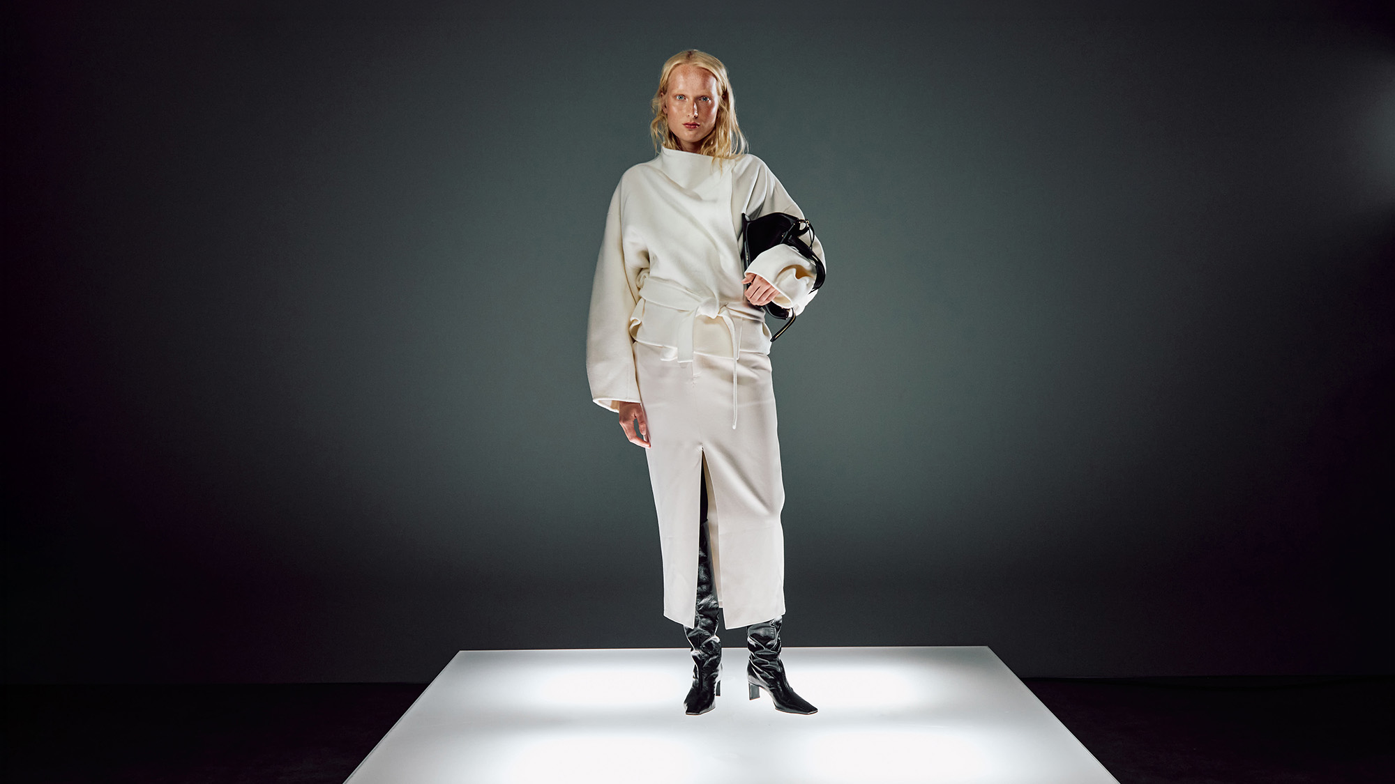Woman wearing cream oversized coat with belt and black boots, holding a black clutch bag