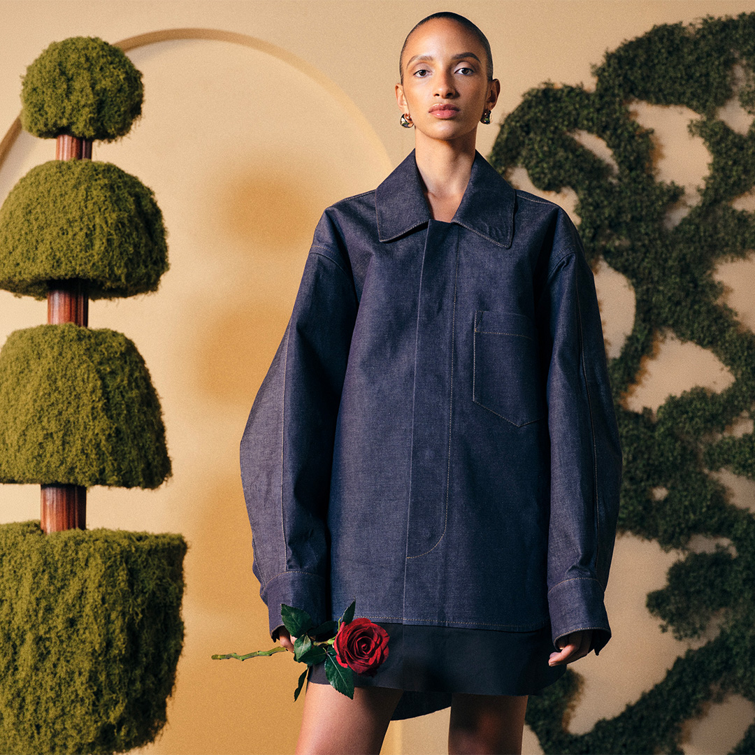 Person in an oversized denim shirt holds a red rose, standing against a backdrop of sculpted greenery.
