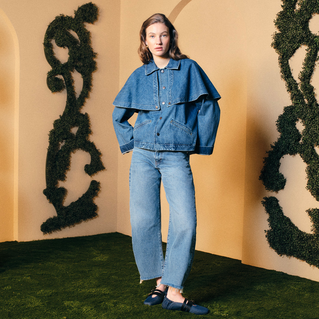 A person wearing a denim outfit stands in a decorative garden with shaped hedges and beige walls.
