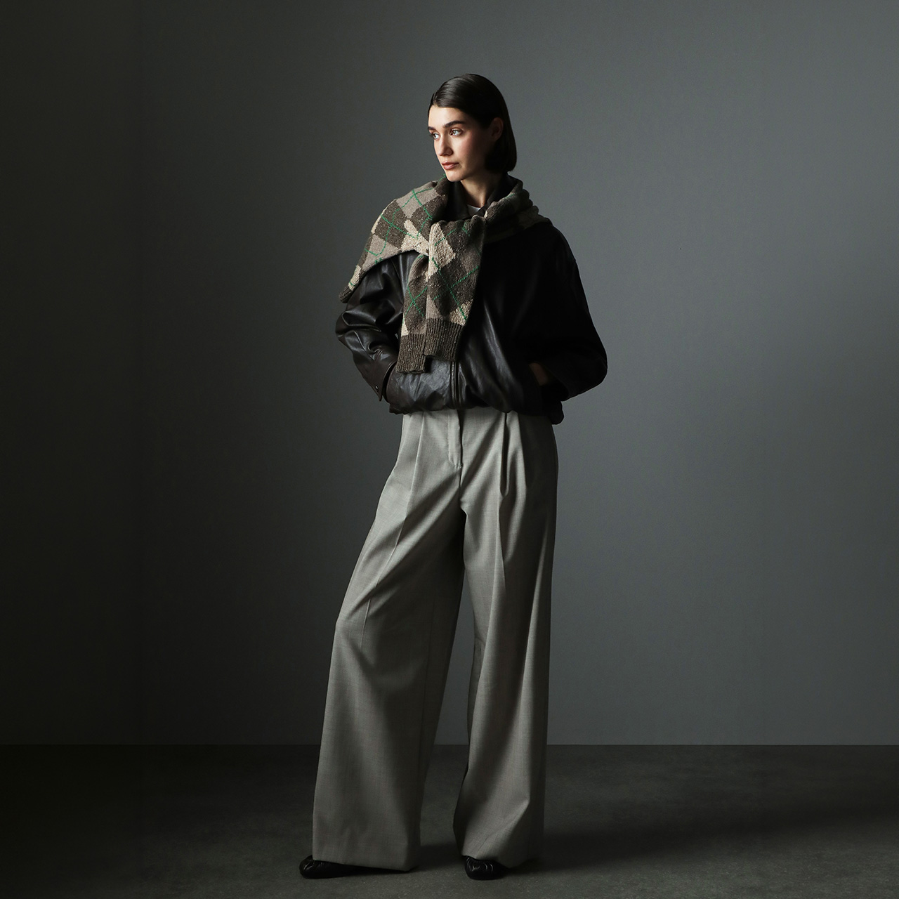 Woman wearing black leather jacket, patterned scarf, and cream-coloured wide-leg trousers against grey background