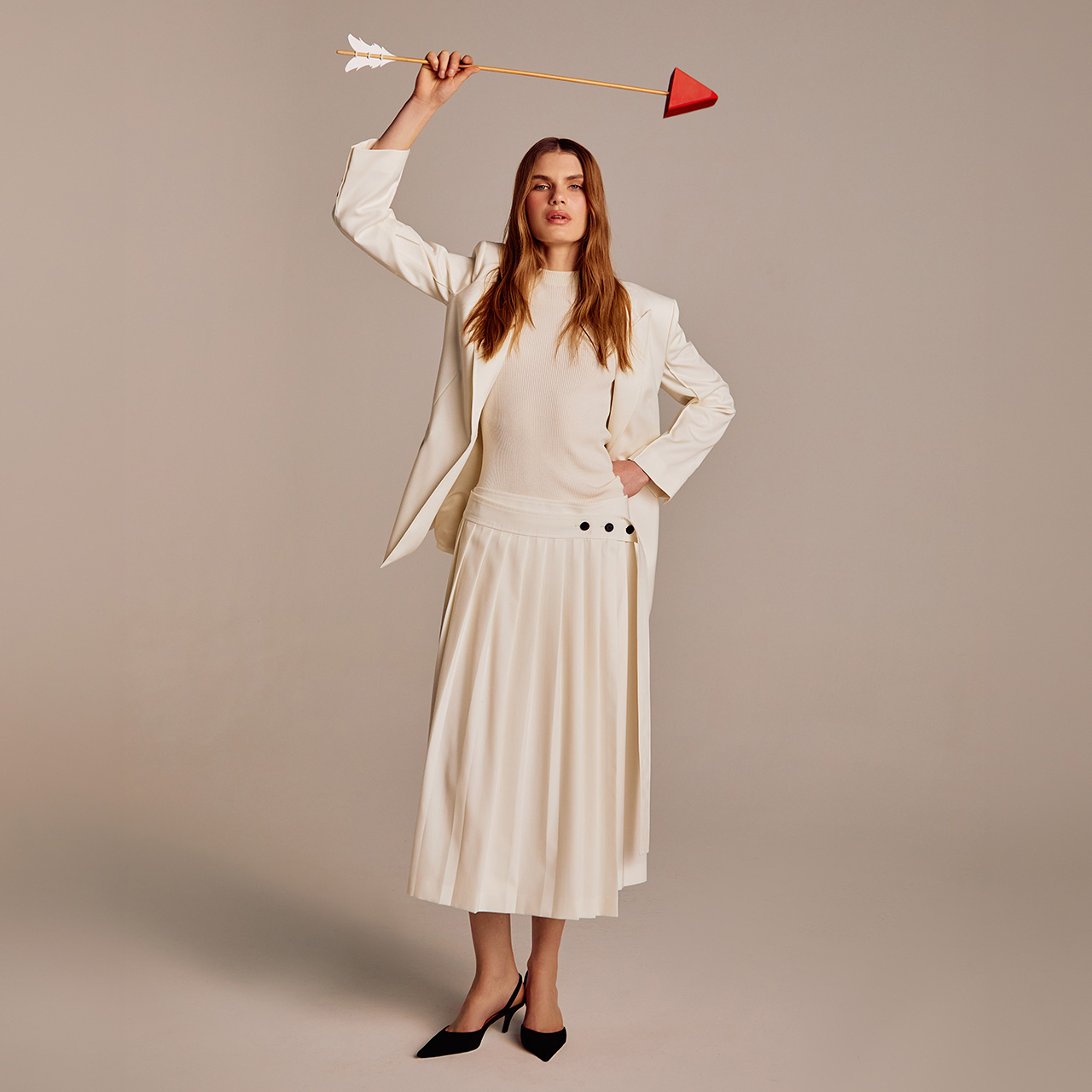 A woman in a white outfit holds an arrow above her head, standing confidently against a neutral background.