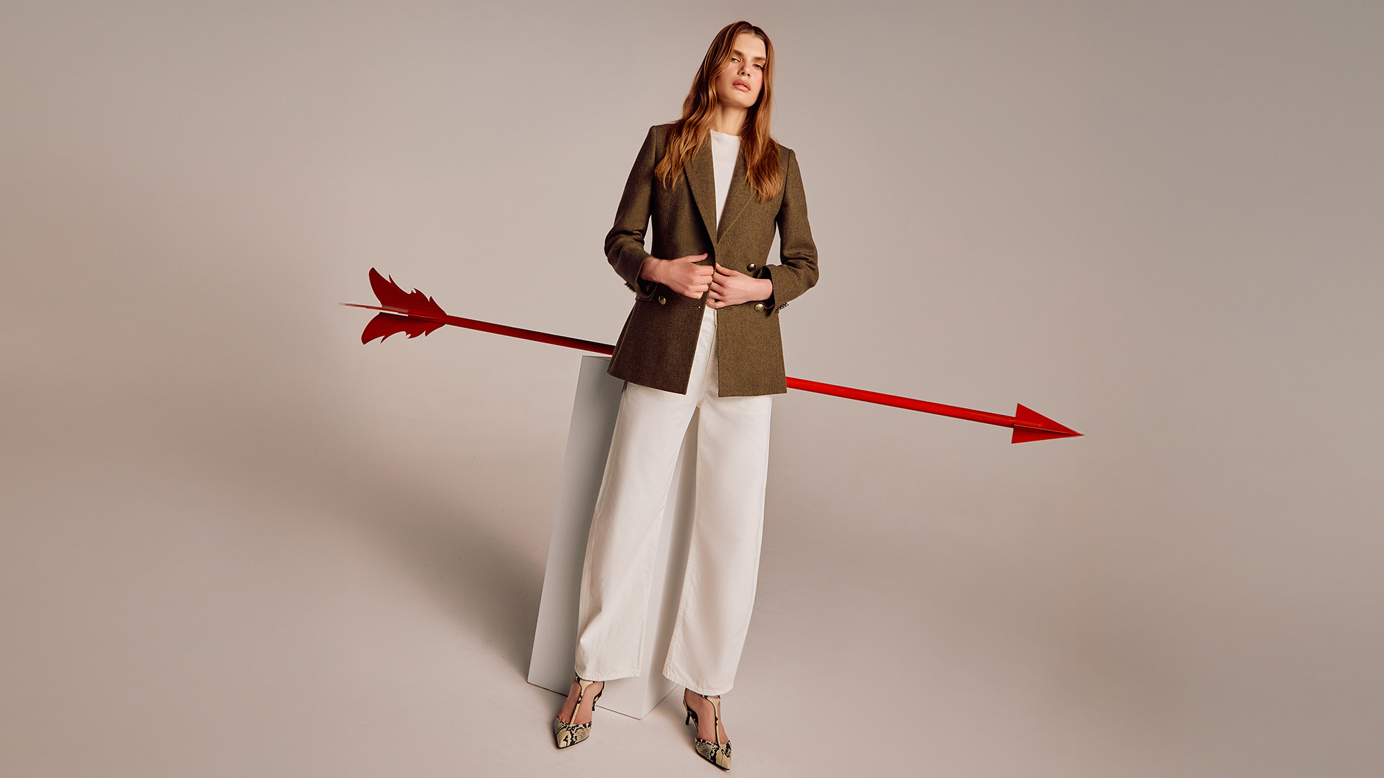 A woman in a blazer and wide-leg pants stands with a large red arrow piercing through her.