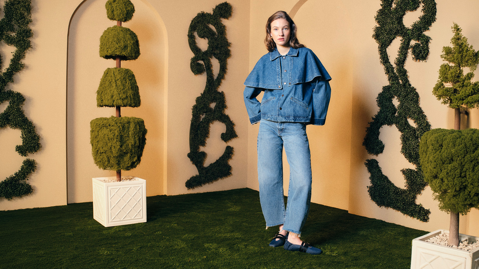 A woman in a flowing yellow dress and sunglasses walks past green hedges, holding a small black purse, against a cloudy blue sky backdrop.