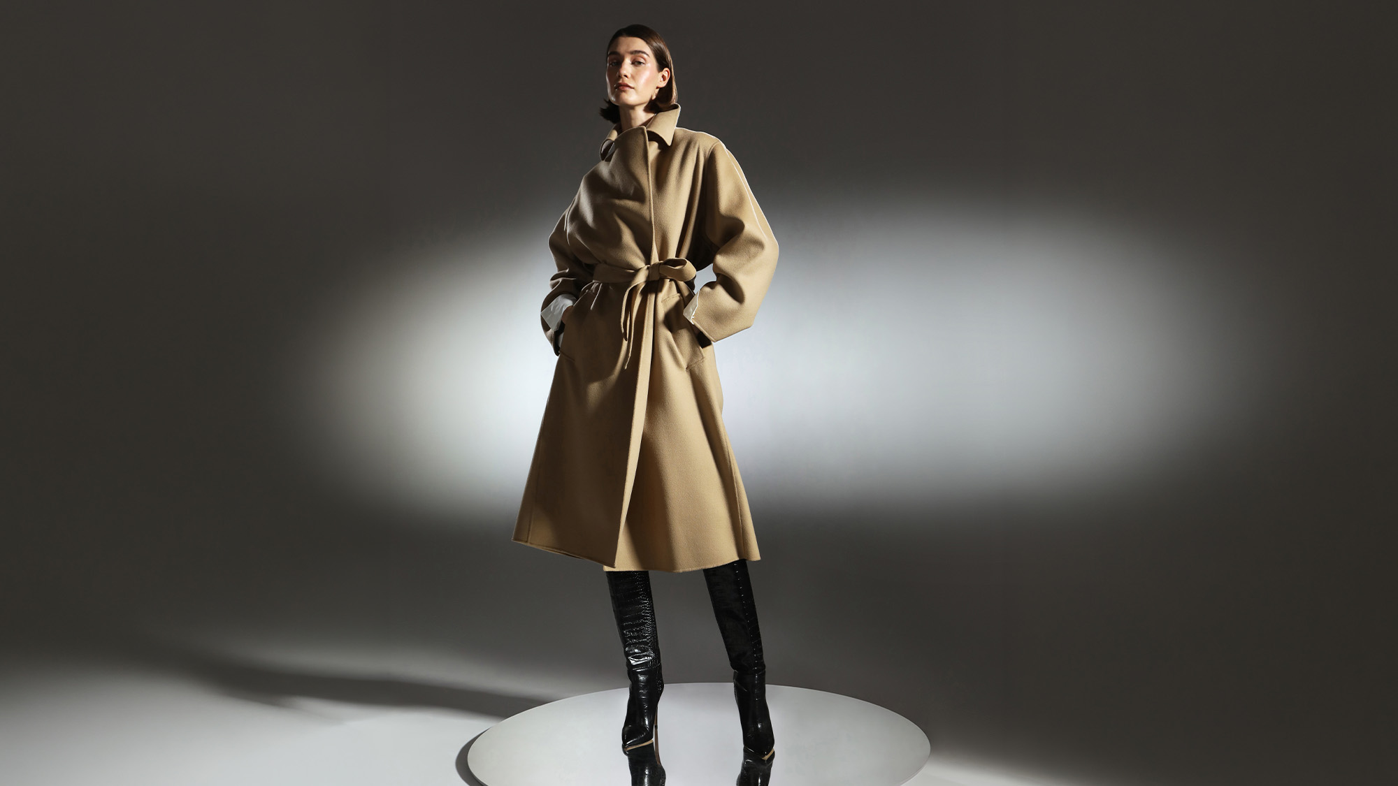 Woman wearing a beige wool coat with tie waist and black leather boots against a neutral backdrop