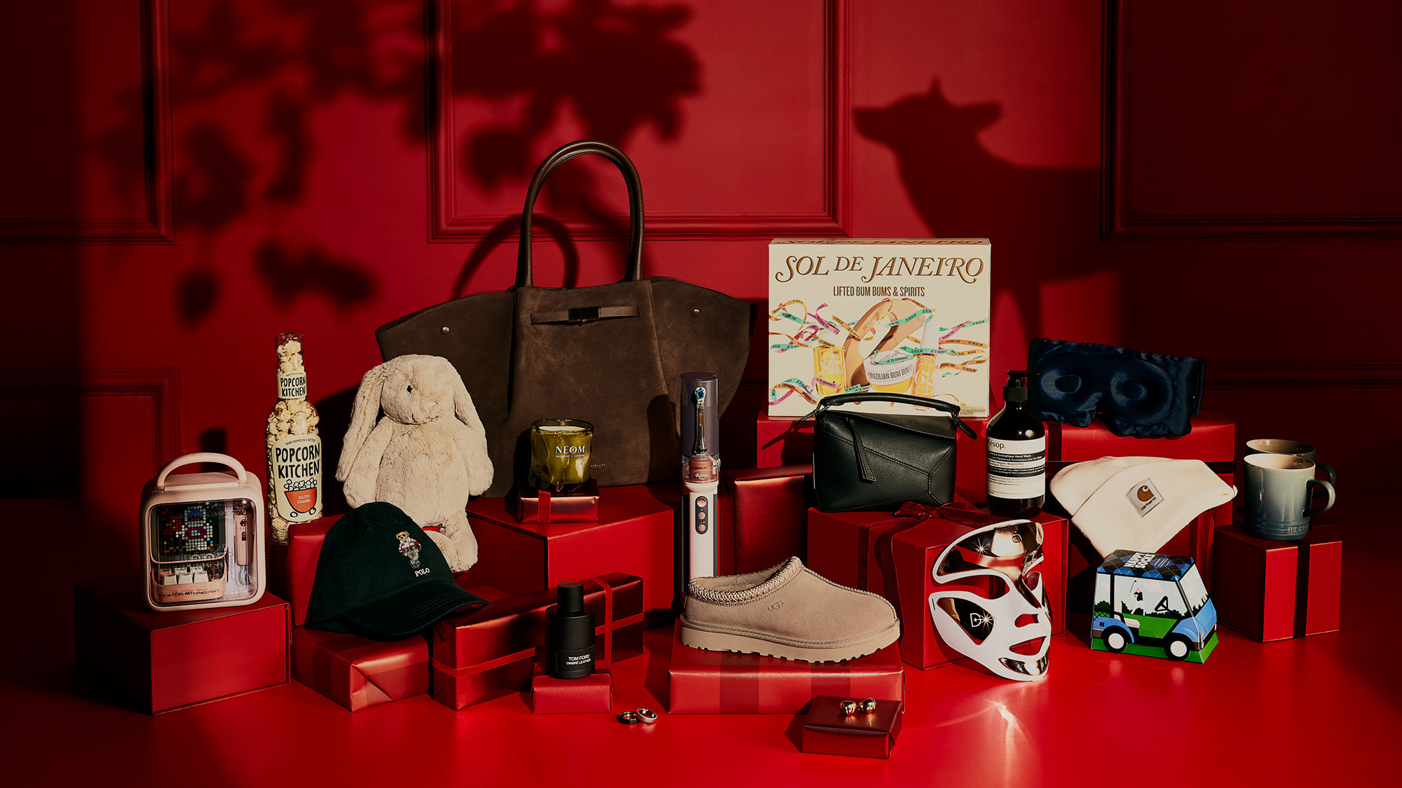 Curated gift collection displayed on red blocks featuring luxury handbag, designer shoes, cosmetics, homeware, and accessories