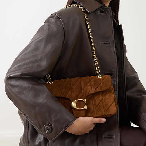 Person in a brown leather coat holding a brown quilted handbag with a gold chain strap and clasp.