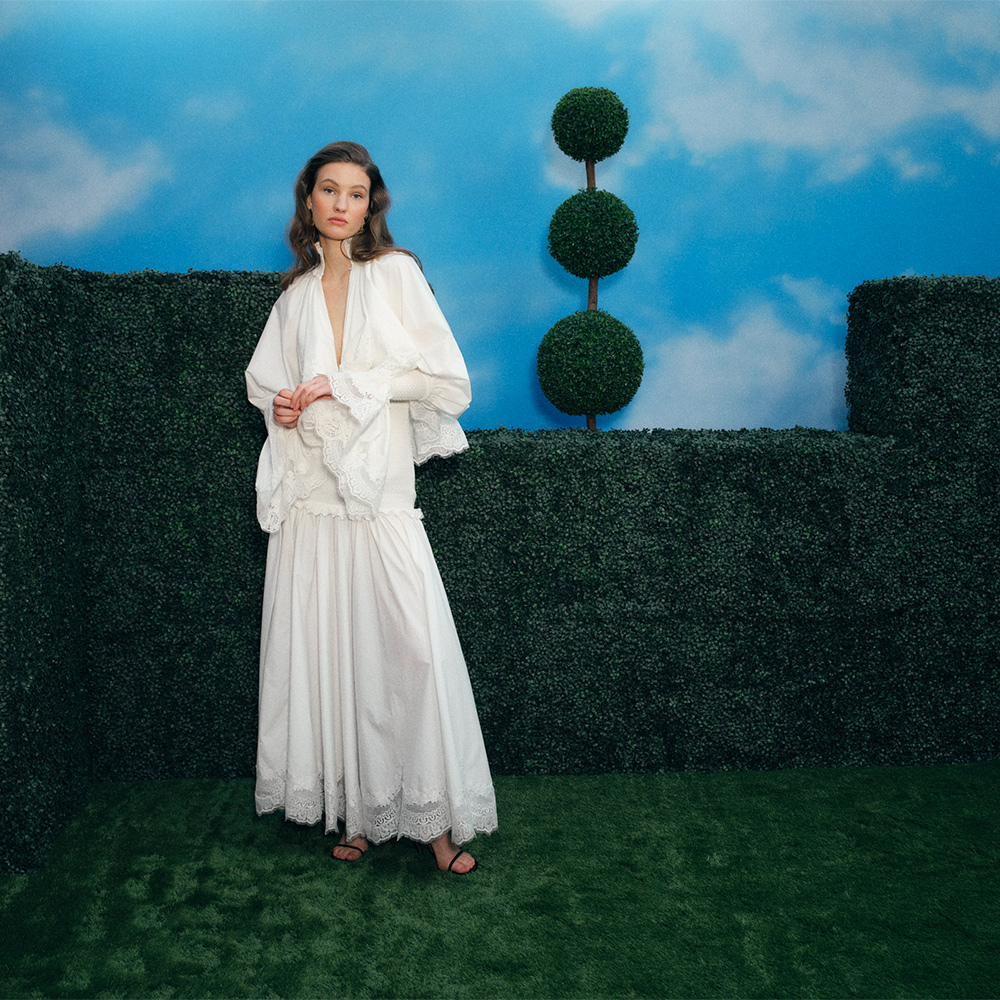 A woman in a flowing white dress stands by manicured hedges and topiary against a backdrop of blue sky with clouds.