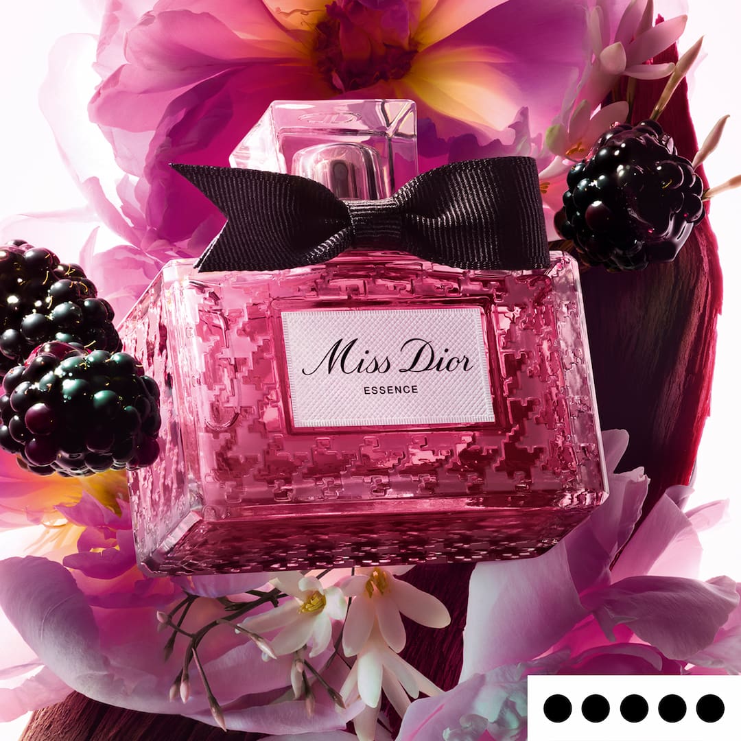 A pink Miss Dior Essence perfume bottle with a black bow, surrounded by flowers and blackberries.
