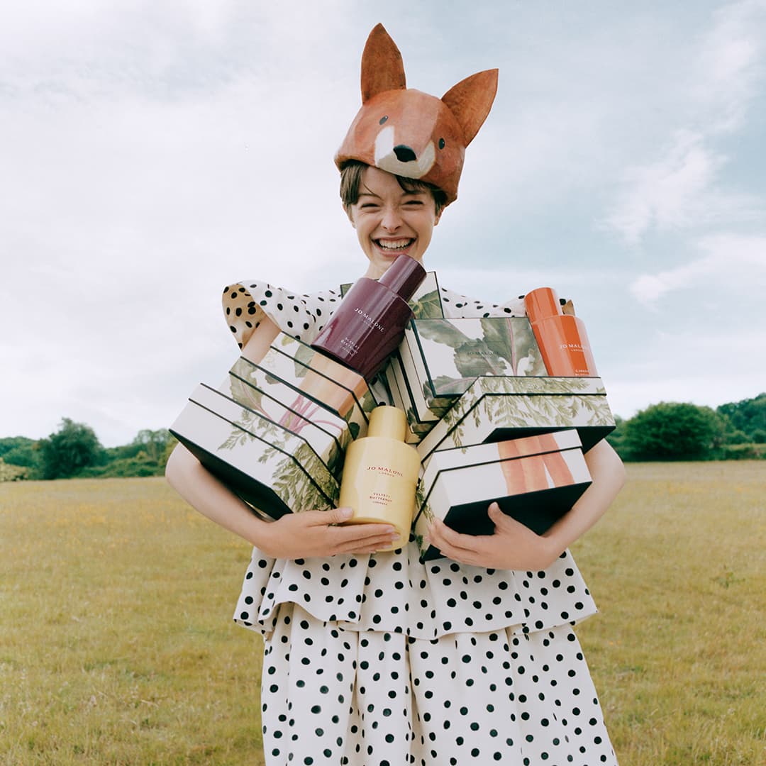 Child wearing fox costume mask holding stacked gift boxes and fragrance bottles in field with countryside background