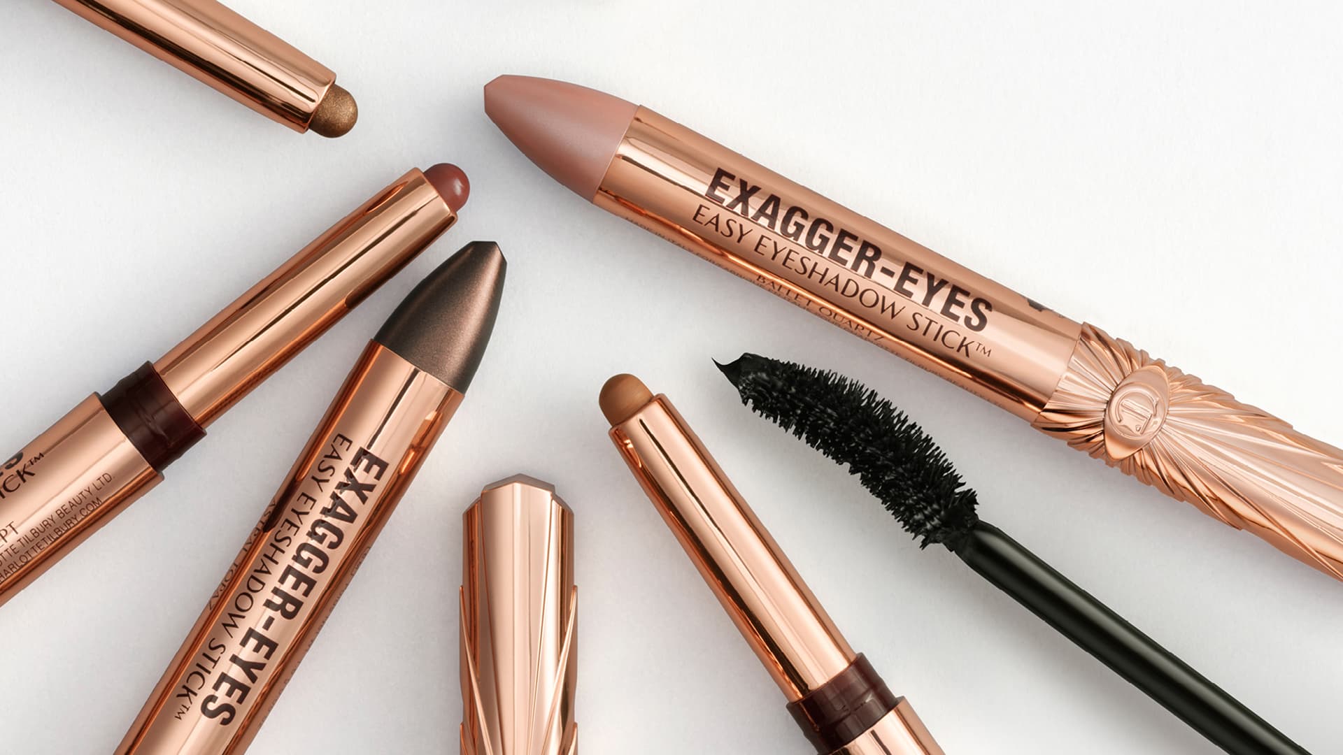Collection of rose-gold eyeshadow sticks, mascara, and lipsticks displayed with product names including Exagger-Eyes Easy Eyeshadow Stick