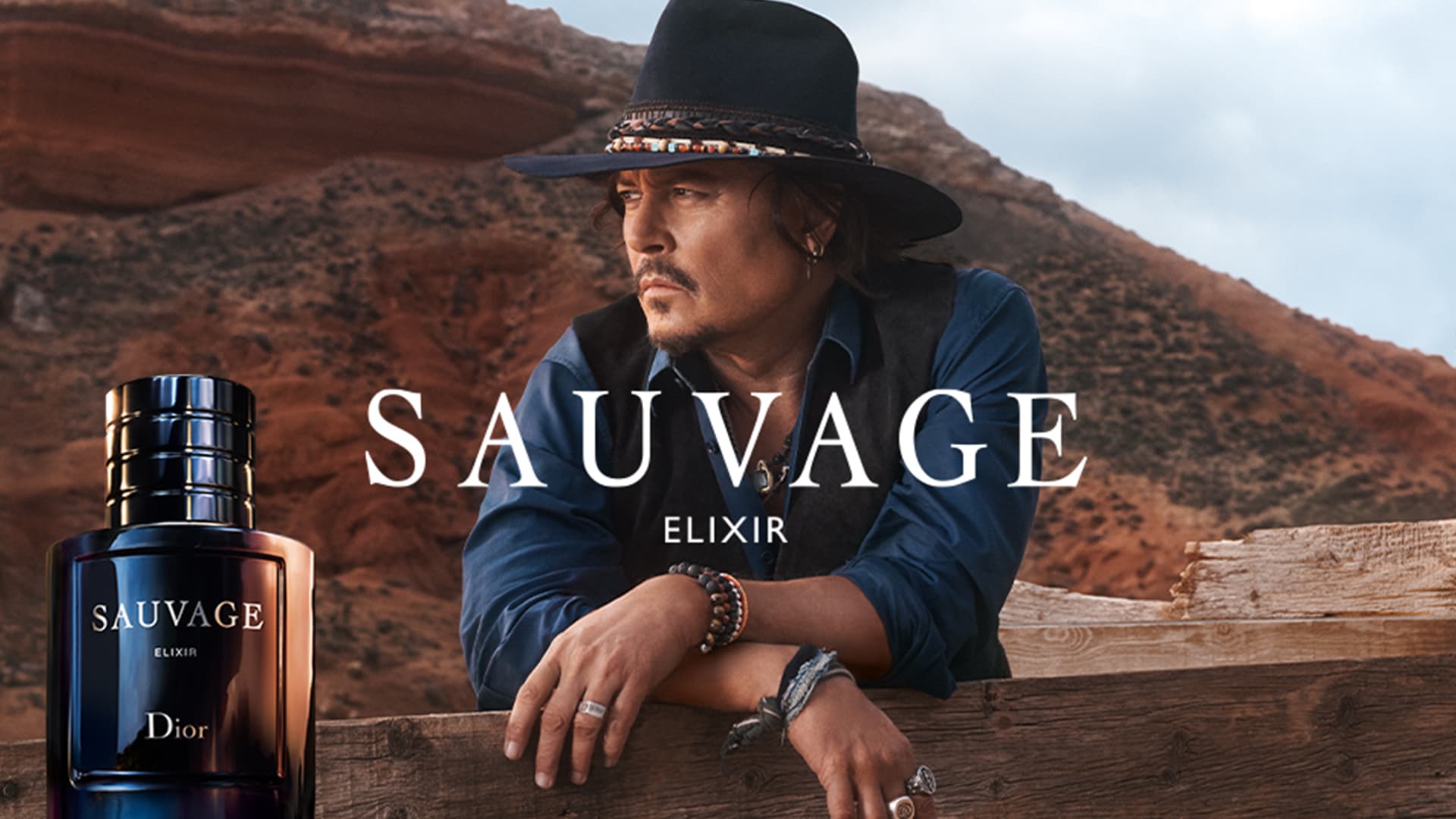 Dior Sauvage Elixir fragrance bottle beside a man wearing a black cowboy hat and blue shirt in desert landscape