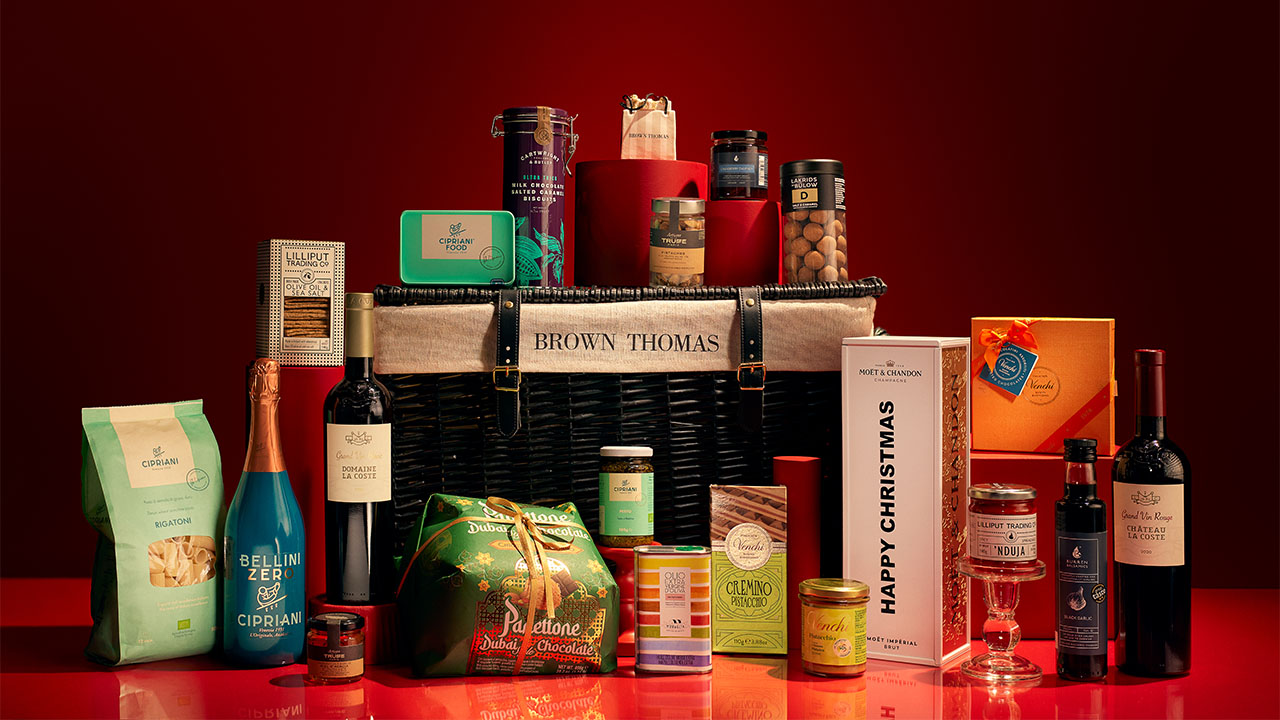 Assortment of gourmet foods and drinks, including wine and chocolates, arranged around a Brown Thomas basket on a red background.