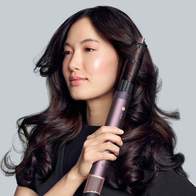 A person with long, wavy hair holds a curling device against a light gray background.