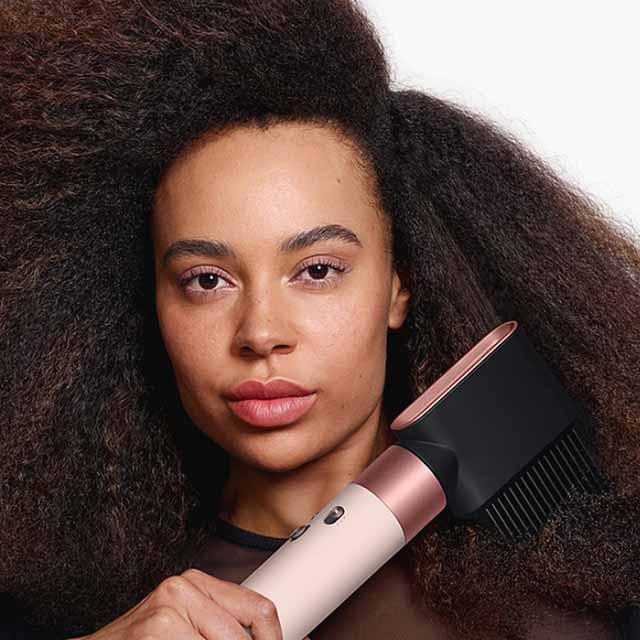 A person holds a hair dryer with a comb attachment next to their voluminous, curly hair.