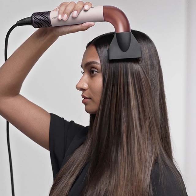 A woman uses a hair styling tool with a curved nozzle on her straight, long hair.