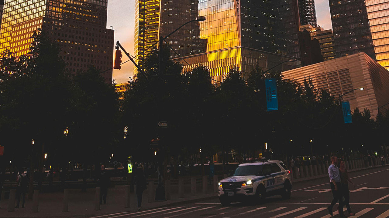 City street at dusk with skyscrapers reflecting golden light, a police car, and pedestrians crossing.
